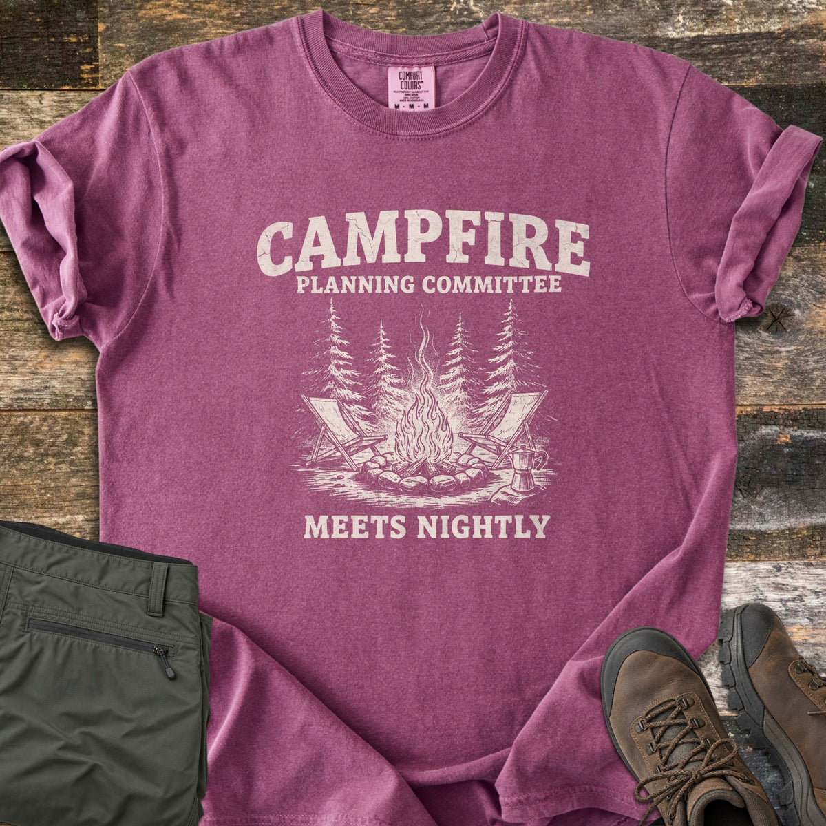 Campfire Planning Committee Comfort Colors T-shirt in Berry, laid flat. Cream print reads Campfire Planning Committee above a sketch of a stone-ringed campfire with two folding chairs, pine trees and a coffee pot, then Meets Nightly. Olive pants and brown hiking boots lie on weathered wood.