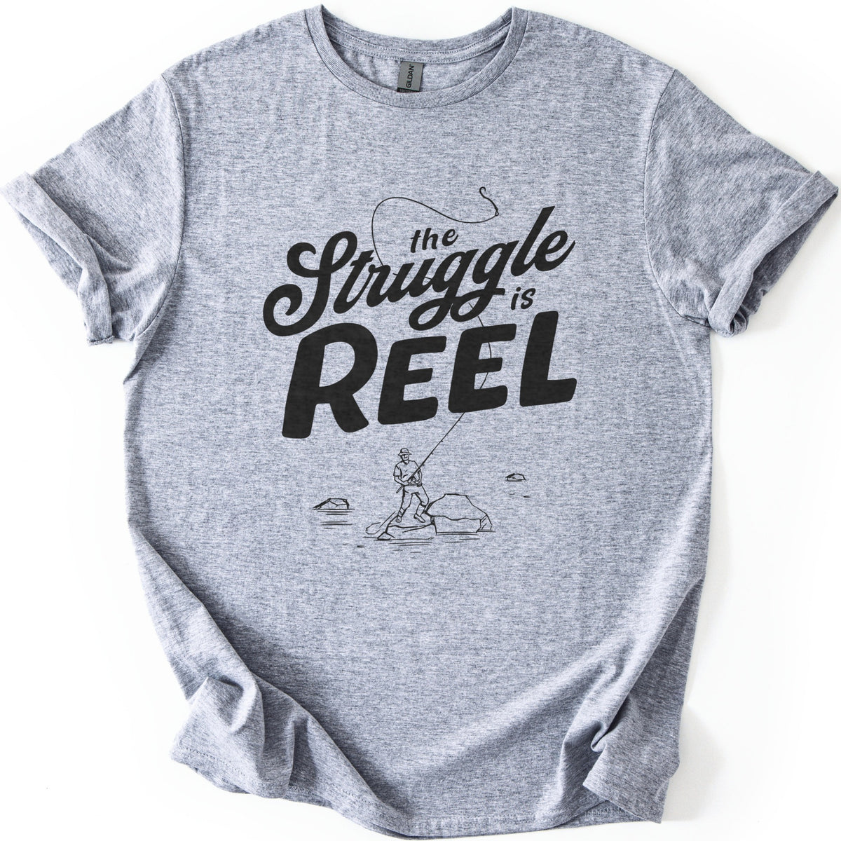 Struggle Is Reel Angler Comfort Colors T-shirt in Sport Grey, laid flat. Black text reads the Struggle is REEL, with a fishing line arcing over the words to a hook. Below, a line drawing of an angler in a hat casts from a rock in the water. Sleeves cuffed, on a white background.