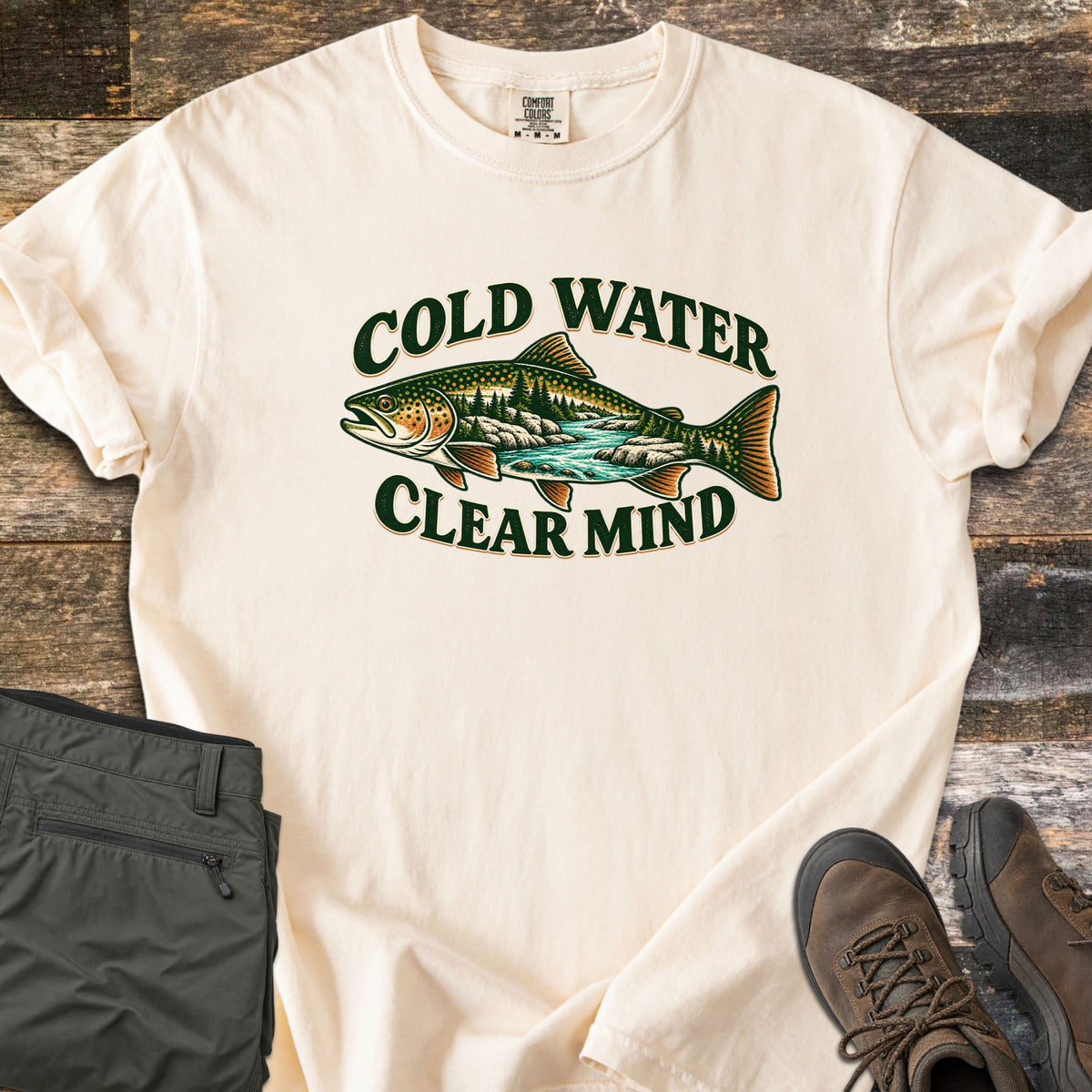 Cold Water Clear Mind Comfort Colors T-shirt in Ivory, laid flat. A speckled trout whose body holds a pine forest and flowing river appears between the arched words Cold Water and Clear Mind. Folded olive pants and brown leather hiking boots rest on weathered wood boards.