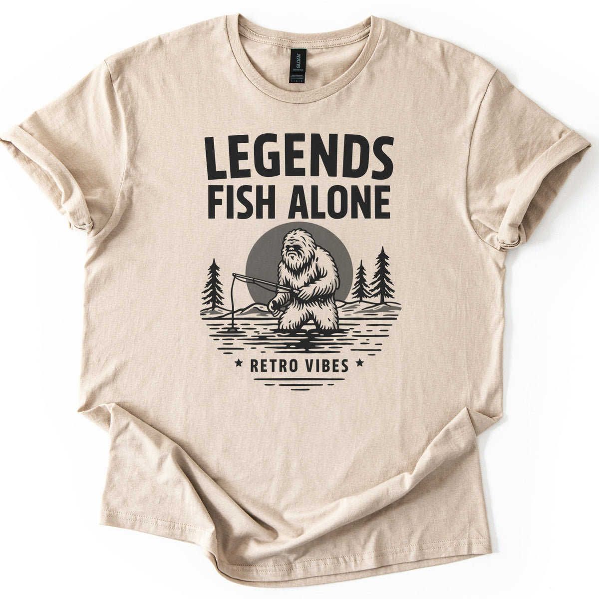 Legends Fish Alone Bigfoot Comfort Colors T-shirt in Sand, laid flat. Black print of Bigfoot fishing knee-deep in a lake before a gray moon and pine trees, under the words Legends Fish Alone and above Retro Vibes between two stars. Sleeves cuffed, on a white background.