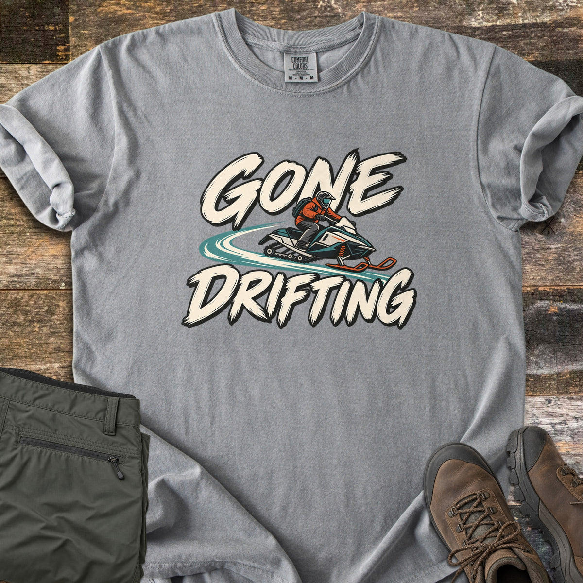 Gone Drifting Comfort Colors T-shirt in Grey, laid flat. Cream brush letters read Gone Drifting around a rider in an orange jacket and helmet on a teal snowmobile with a swirling snow trail. On weathered wood beside folded olive pants and brown hiking boots.