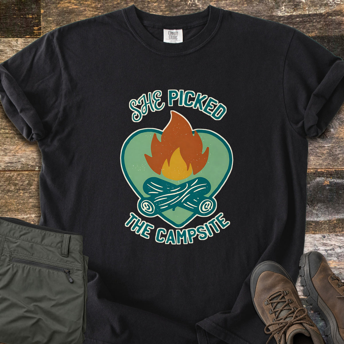 She Picked The Campsite Comfort Colors T-shirt in Black, laid flat. A campfire with crossed logs sits on a green heart, with the words She Picked above and The Campsite below. Sleeves are rolled on a rustic wood floor beside folded olive hiking pants and brown hiking boots.
