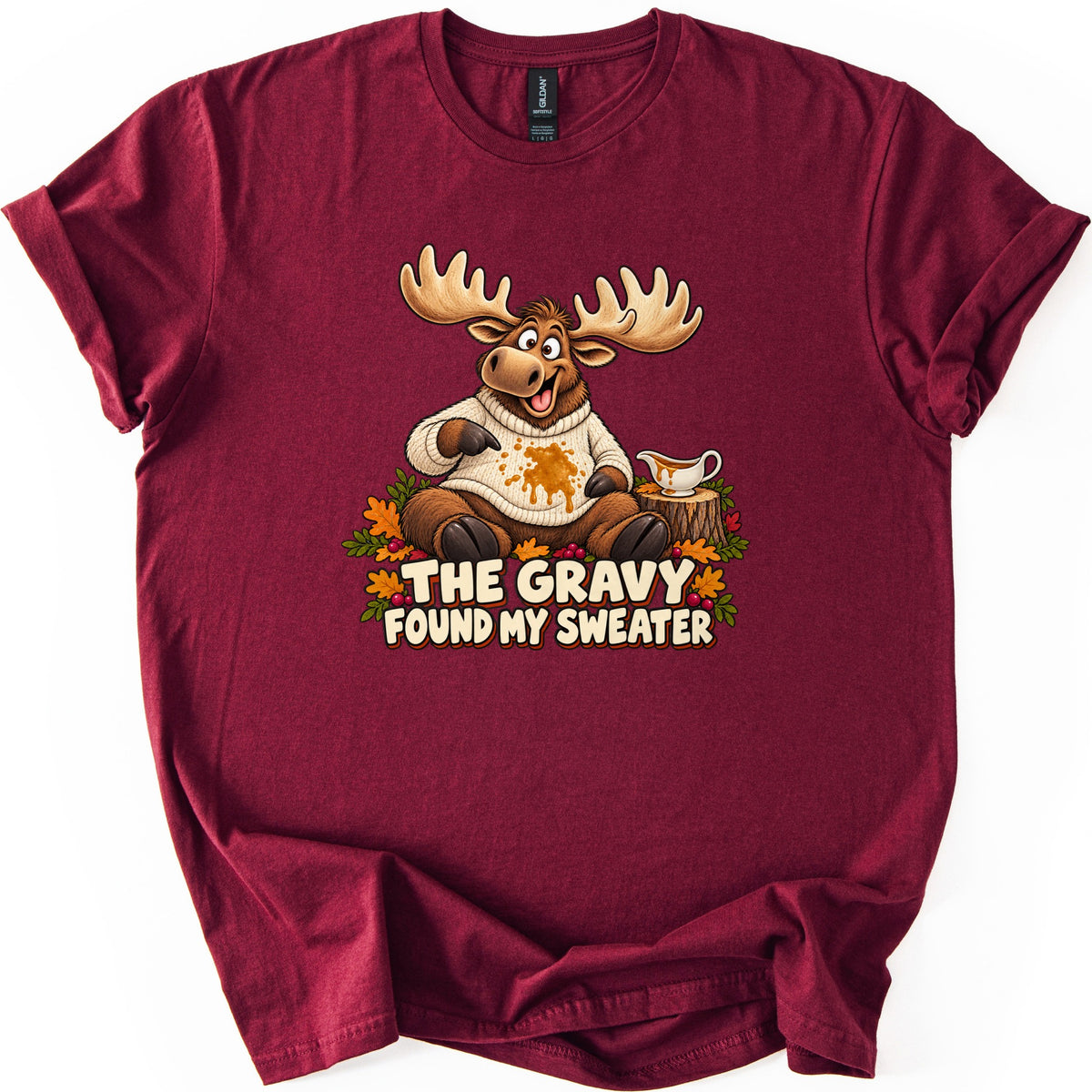 The Gravy Found My Sweater Comfort Colors T-shirt in Maroon, laid flat. A cartoon moose with large antlers points at a gravy stain on his cream sweater, beside a gravy boat on a tree stump and autumn leaves, above the words The Gravy Found My Sweater. Sleeves are rolled on white.