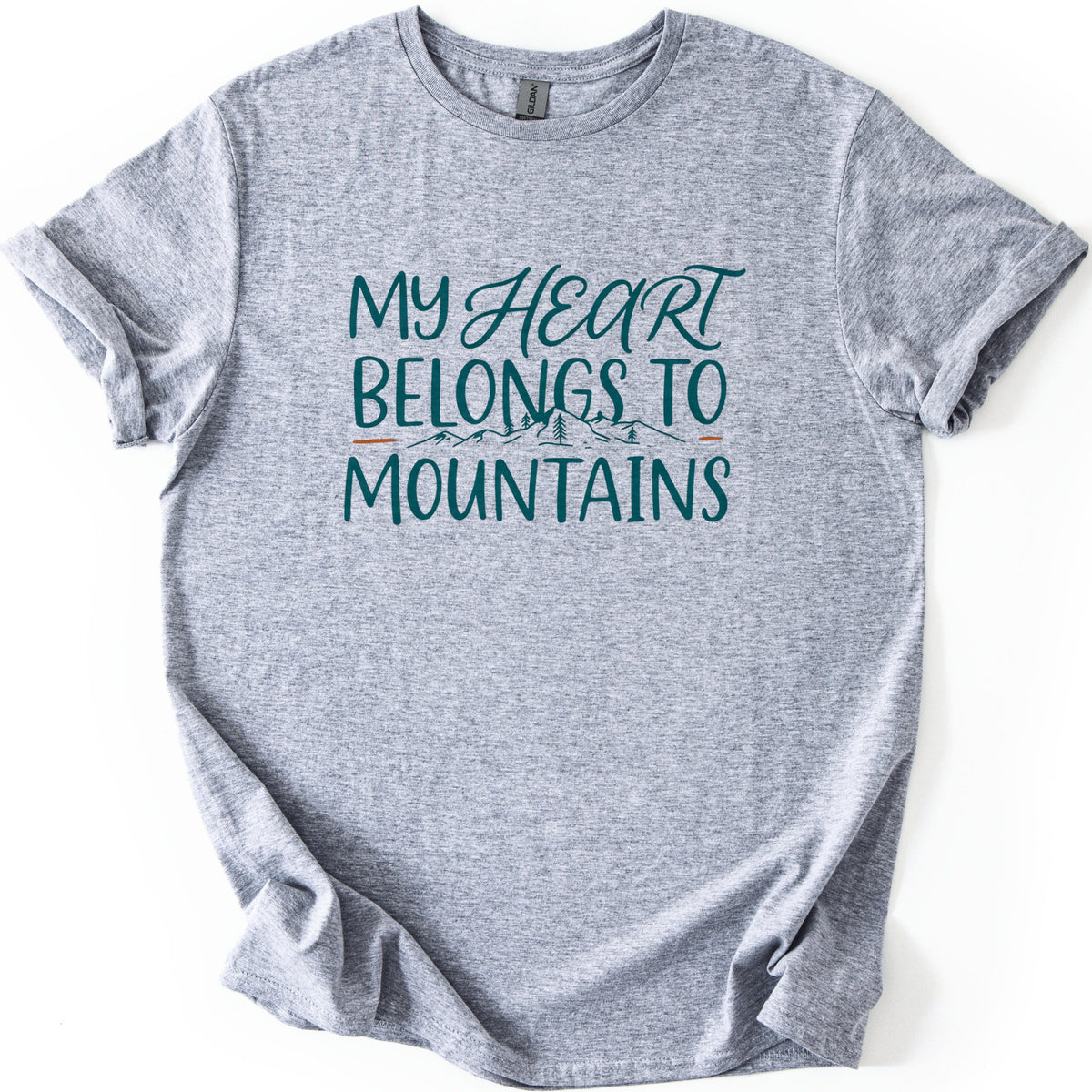 My Heart Belongs to Mountains Comfort Colors T-shirt in Sport Grey, laid flat. Teal lettering reads My Heart Belongs to Mountains, with Heart in script above a small line drawing of mountains and pine trees between orange dashes. The shirt has cuffed sleeves and lies on a white background.