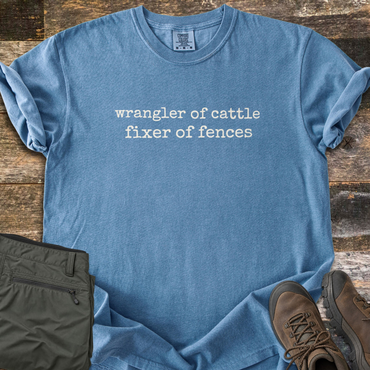 Fixer of Fences Comfort Colors T-shirt in Blue Jean, laid flat. Off-white typewriter-style text across the chest reads wrangler of cattle, fixer of fences. The shirt has rolled sleeves and lies on weathered wood planks beside folded olive green pants and brown hiking boots.