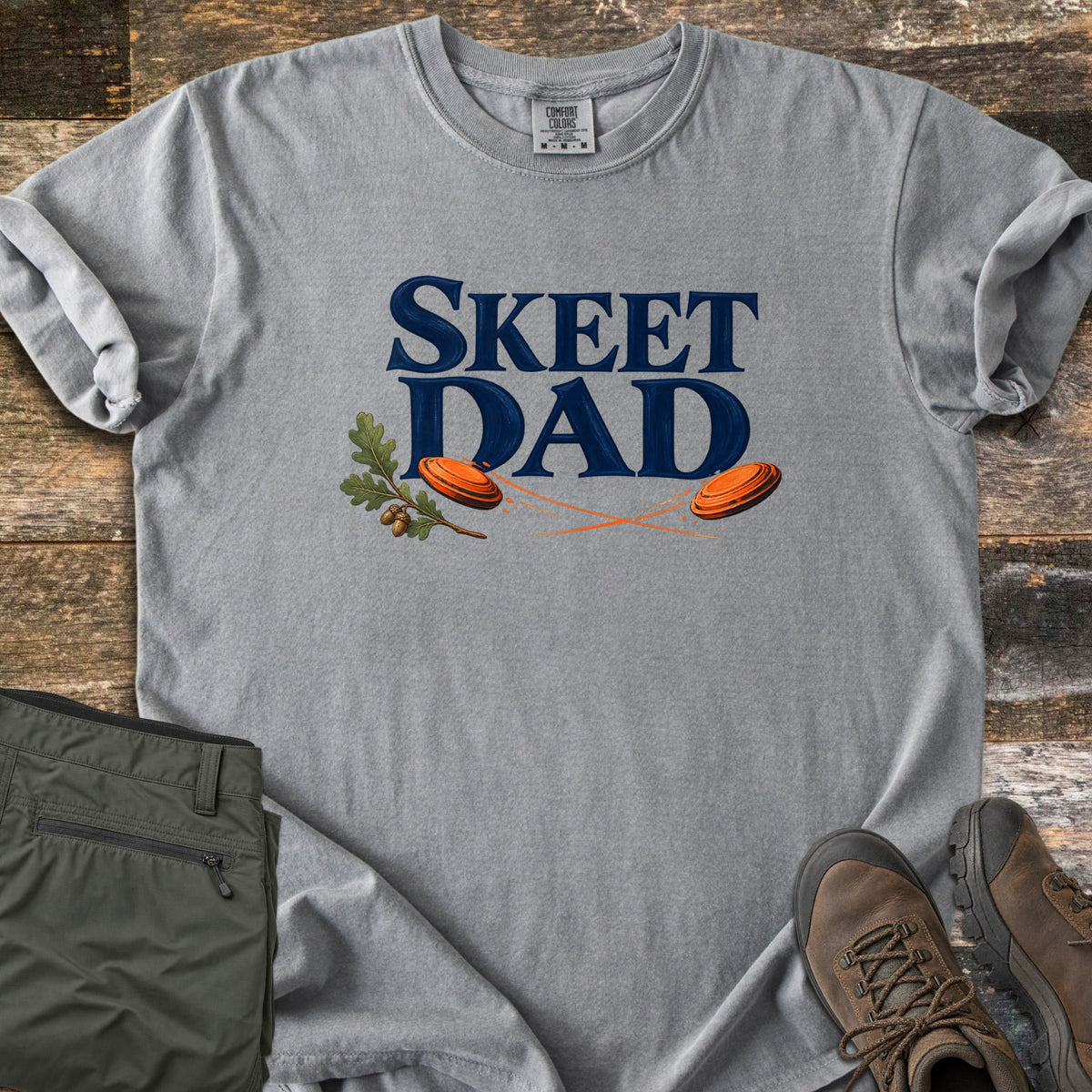Skeet Dad Comfort Colors T-shirt in Grey, laid flat. Navy text reads Skeet Dad above two orange clay targets with motion trails and an oak sprig with acorns. Sleeves are rolled on a wood floor beside folded olive cargo pants and brown hiking boots.