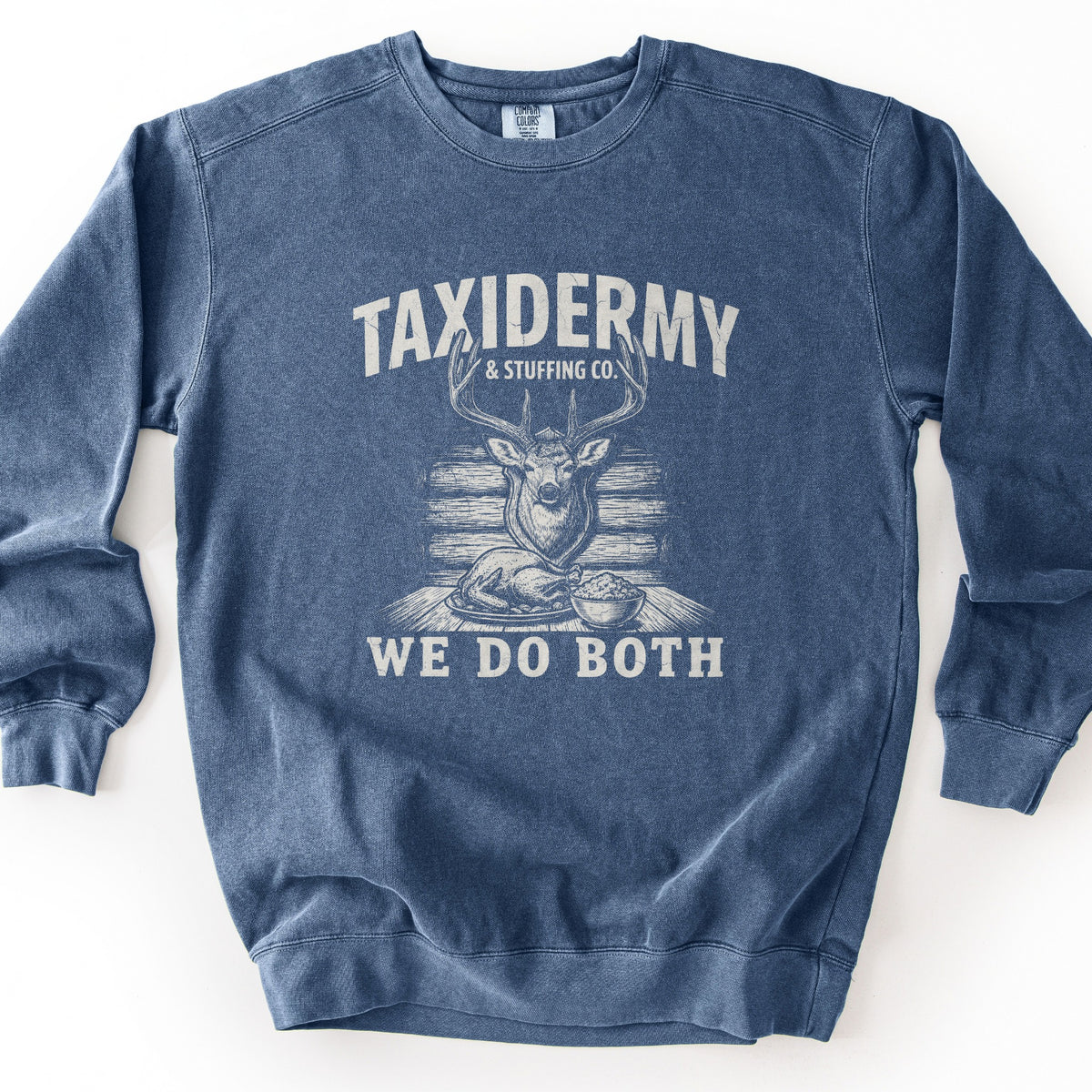 Taxidermy & Stuffing Co. We Do Both Comfort Colors crewneck sweatshirt in Denim, laid flat. A distressed cream print shows a mounted deer head above a roast turkey and a bowl of stuffing on a table, with the words Taxidermy & Stuffing Co. above and We Do Both below. Shown on a white background.