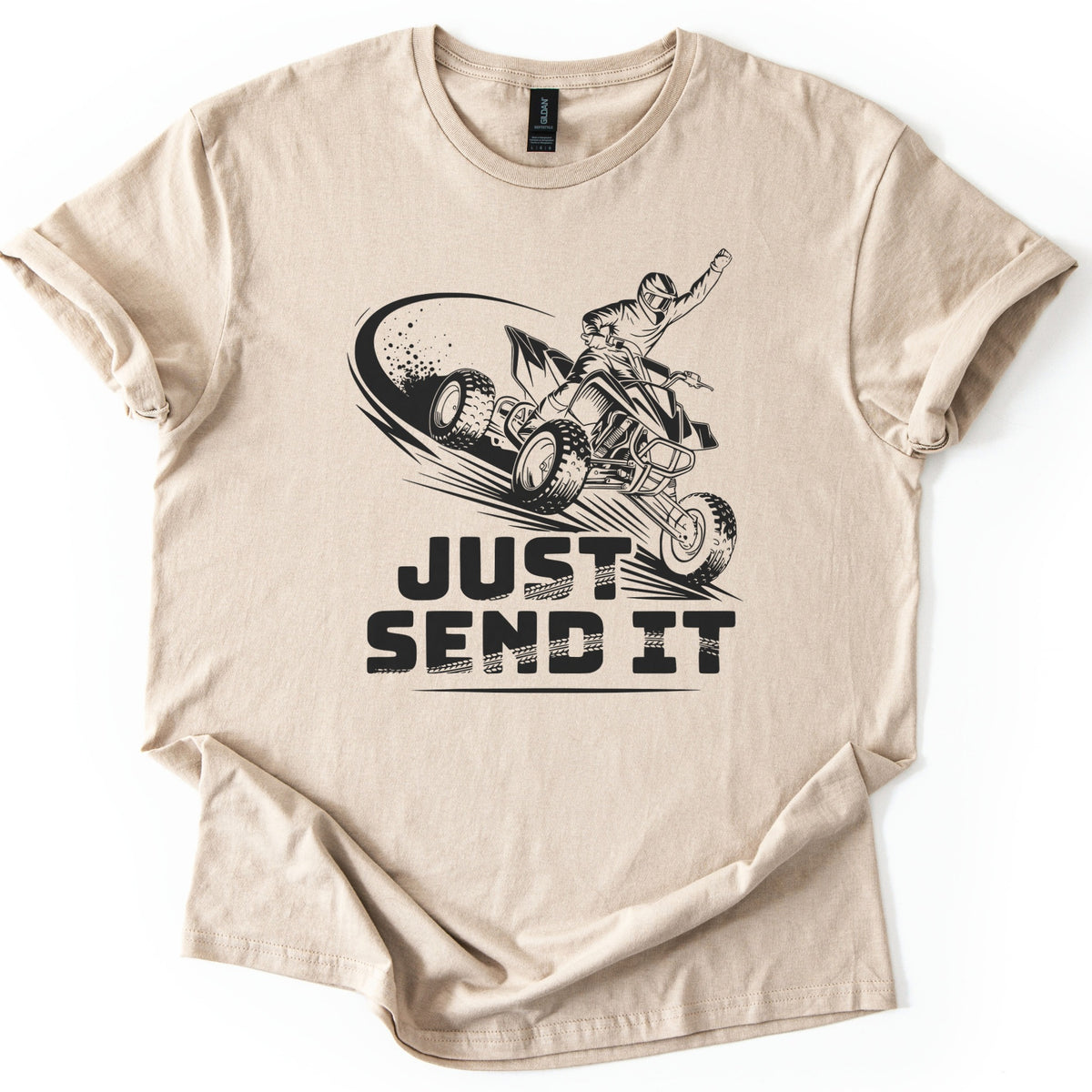 ATV Just Send It Comfort Colors T-shirt in Sand, laid flat. A black illustration shows a helmeted rider on a quad ATV mid-jump, fist raised, with a swoosh of dust, above the words Just Send It in bold tire-marked letters. The shirt lies on a white background.