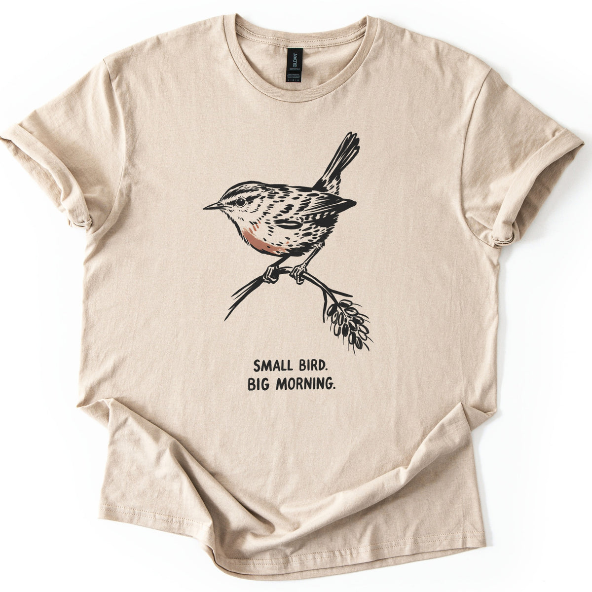 Small Bird Big Morning Comfort Colors T-shirt in Sand, laid flat. A black ink sketch of a wren with a rust-colored breast perches on a twig with a seed head, above the words Small Bird. Big Morning. The sleeves are rolled up, on a white background.