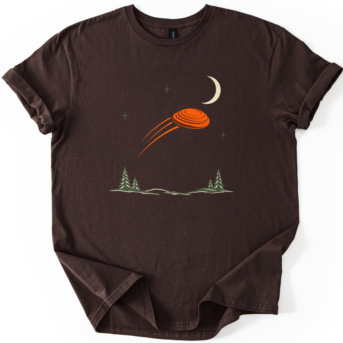 Moonrise Clay Comfort Colors T-shirt in Dark Chocolate, laid flat. An orange clay target streaks across a night sky with a cream crescent moon and small green stars, above pine trees and low hills. No words are printed. Sleeves rolled, on a white background.