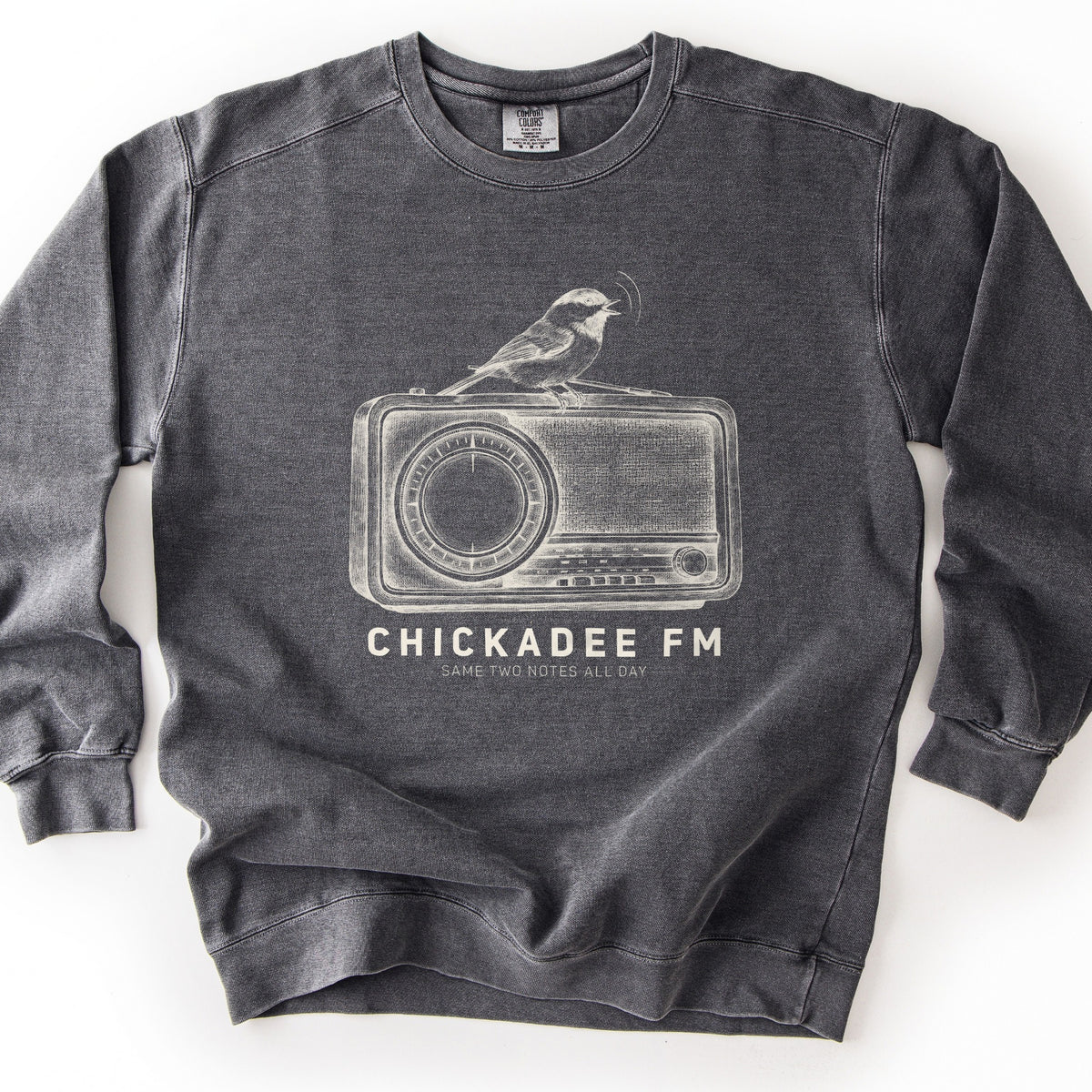 Chickadee FM Comfort Colors crewneck sweatshirt in Pepper, laid flat. A sketched chickadee sings atop a vintage radio, printed in cream above the words CHICKADEE FM and SAME TWO NOTES ALL DAY. It lies on a white surface with the sleeves out to the sides.