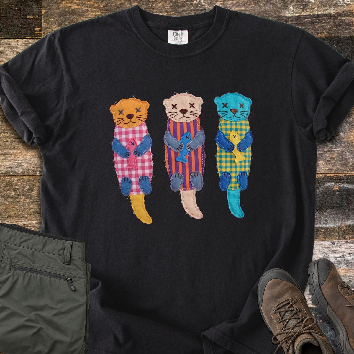 Patchwork Otter Comfort Colors T-shirt in Black, laid flat. Three patchwork otters in orange, peach and turquoise float on their backs, each in a gingham or striped body hugging a small fish, with X-stitched eyes. Olive pants and brown hiking boots sit on a wood floor.