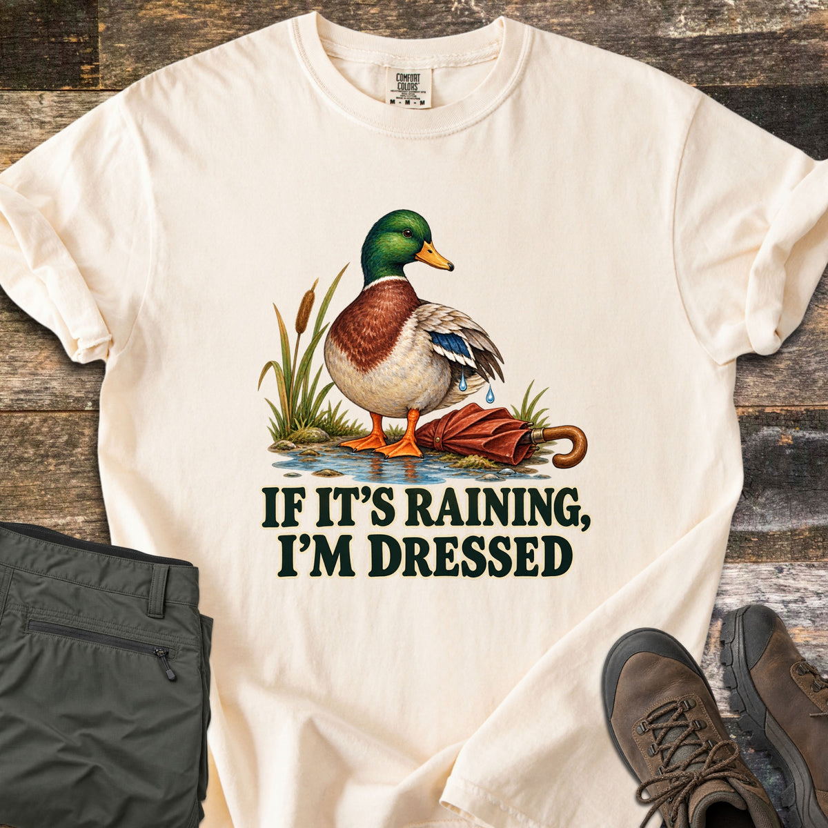 If It's Raining, I'm Dressed Comfort Colors T-shirt in Ivory, laid flat. A mallard duck stands in a puddle by cattails, water dripping off its wing, next to a closed red umbrella, above the words If It's Raining, I'm Dressed in dark green. Olive pants and brown hiking boots lie on weathered wood.