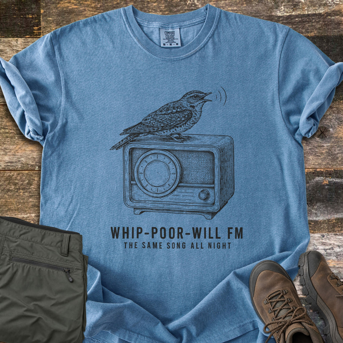 Whip-Poor-Will FM Comfort Colors T-shirt in Blue Jean, laid flat. A sketched whip-poor-will sings atop a vintage radio above the words Whip-Poor-Will FM, The Same Song All Night. It lies with rolled sleeves on weathered wood beside folded olive pants and brown hiking boots.