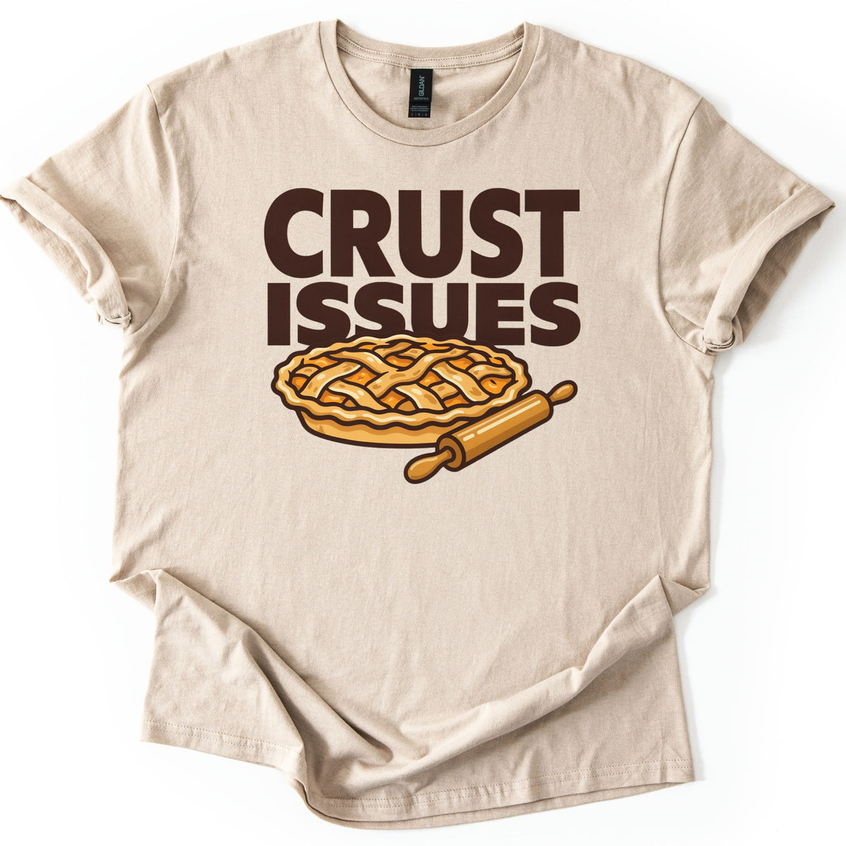 Crust Issues Comfort Colors T-shirt in Sand, laid flat. Dark brown block letters read CRUST ISSUES above a cartoon lattice-top pie with a wooden rolling pin. The shirt has rolled sleeves and lies on a plain white background.