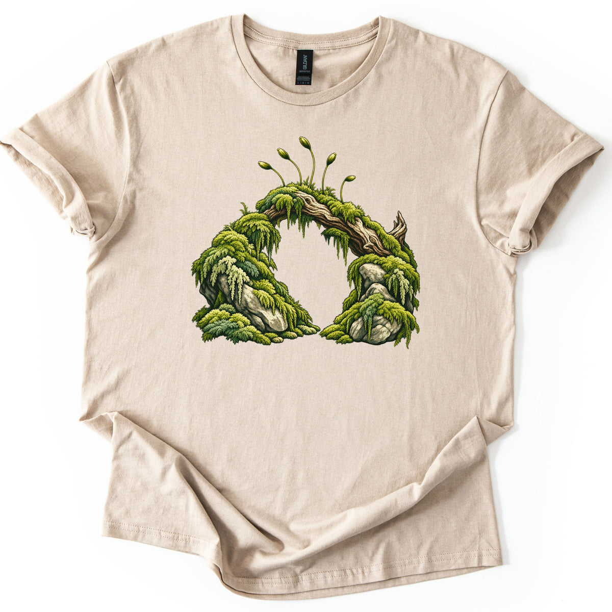 Moss Arch Comfort Colors T-shirt in Sand, laid flat. A fallen log thick with hanging green moss forms an arch over two mossy rocks, topped by slender spore stalks. No words are printed. The sleeves are cuffed and the shirt rests on a white background.