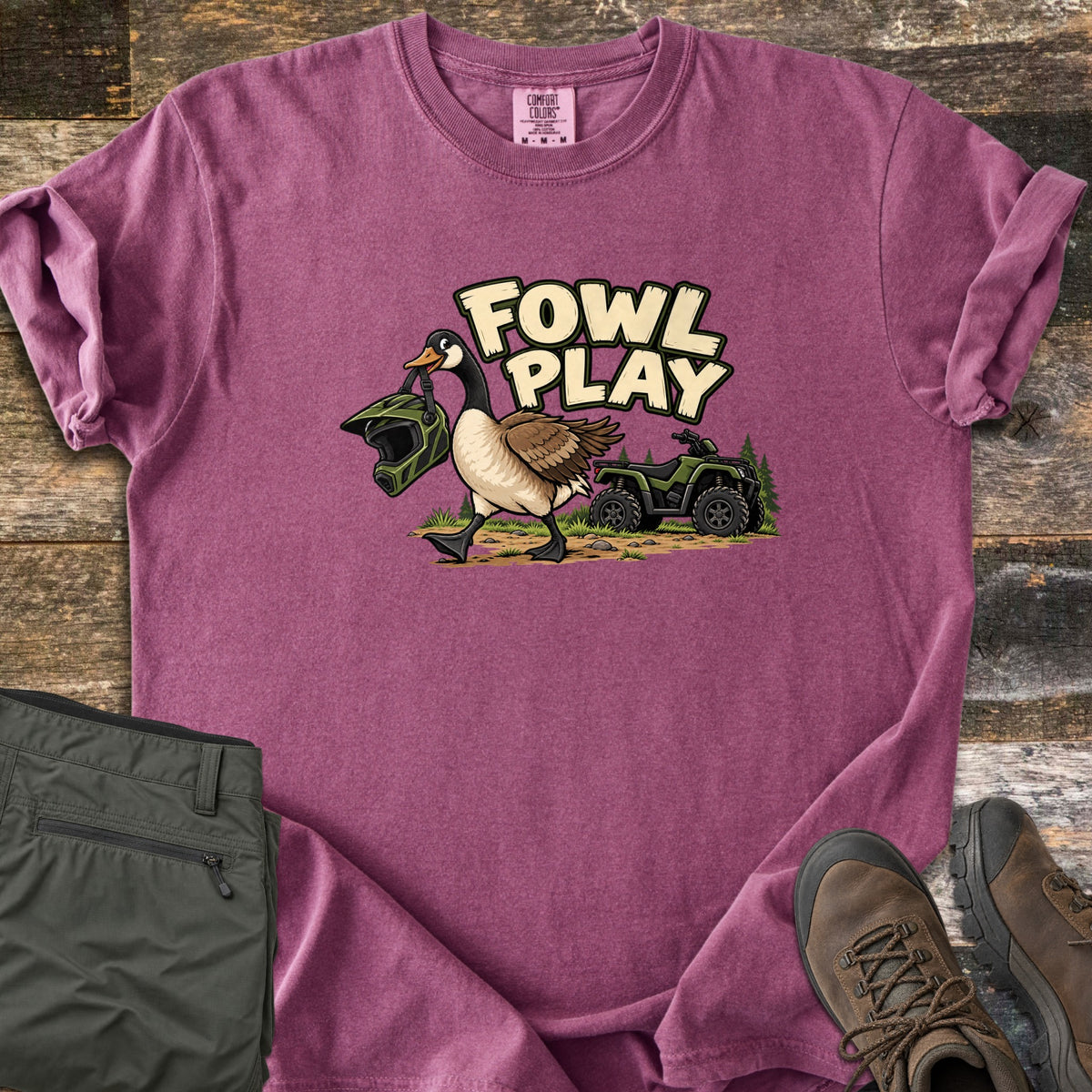 Fowl Play Comfort Colors T-shirt in Berry, laid flat. A cartoon Canada goose carries a green off-road helmet in its beak past a green ATV and pine trees, under the words Fowl Play. Sleeves rolled, on weathered wood with olive pants and brown hiking boots.