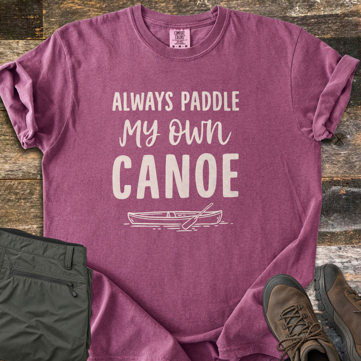 Paddle My Own Canoe Comfort Colors T-shirt in Berry, laid flat. Cream text reads Always Paddle My Own Canoe above a line drawing of a canoe with a paddle on the water. It lies on weathered wood with rolled sleeves, folded olive hiking pants, and brown hiking boots.