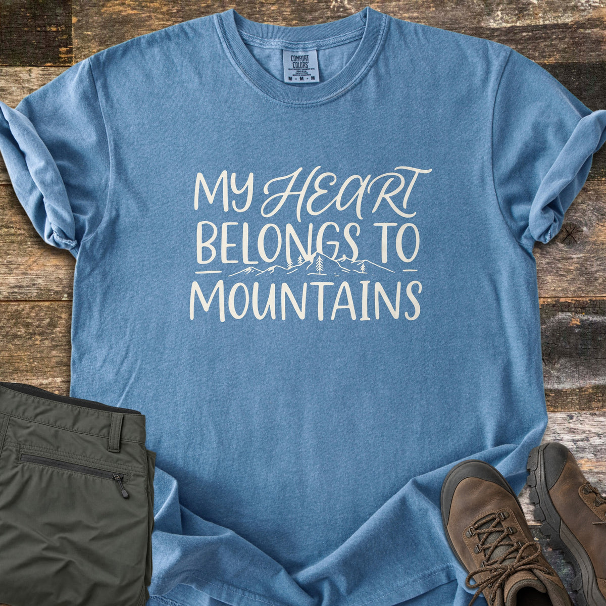 My Heart Belongs to Mountains Comfort Colors T-shirt in Blue Jean, laid flat. Cream lettering reads My Heart Belongs to Mountains, with Heart in script over a sketched range of peaks and pines. Folded olive hiking pants and brown hiking boots lie beside it on rustic wood planks.