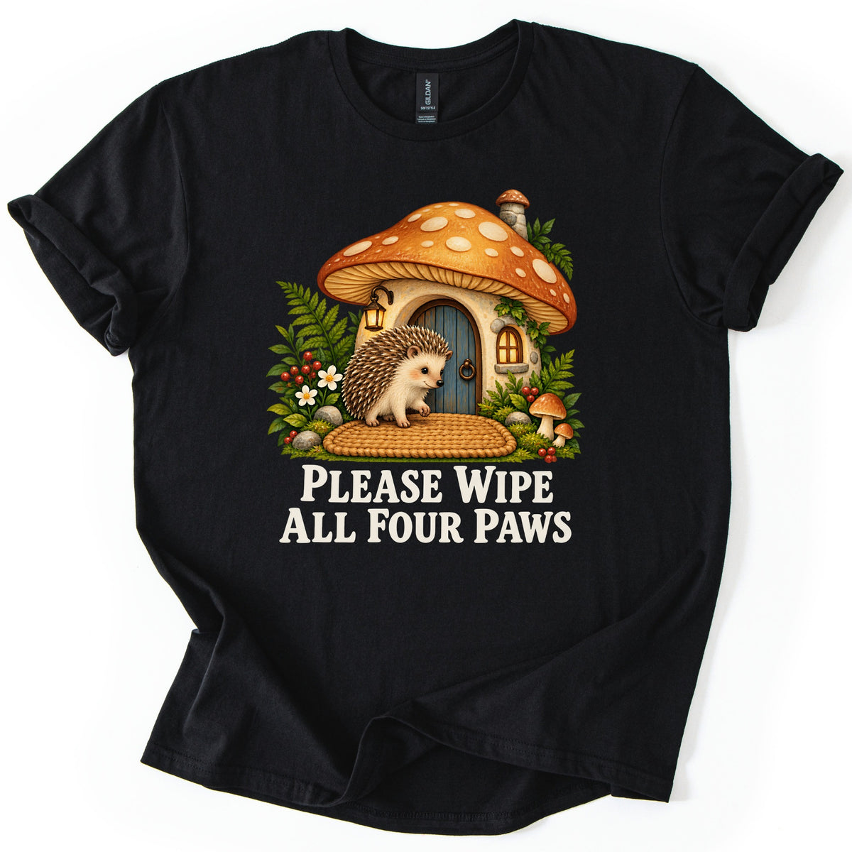 Please Wipe All Four Paws Comfort Colors T-shirt in Black, laid flat. A hedgehog stands on a woven doormat outside a mushroom cottage with an orange spotted cap, lantern and blue door, framed by ferns. Below, cream text reads Please Wipe All Four Paws.