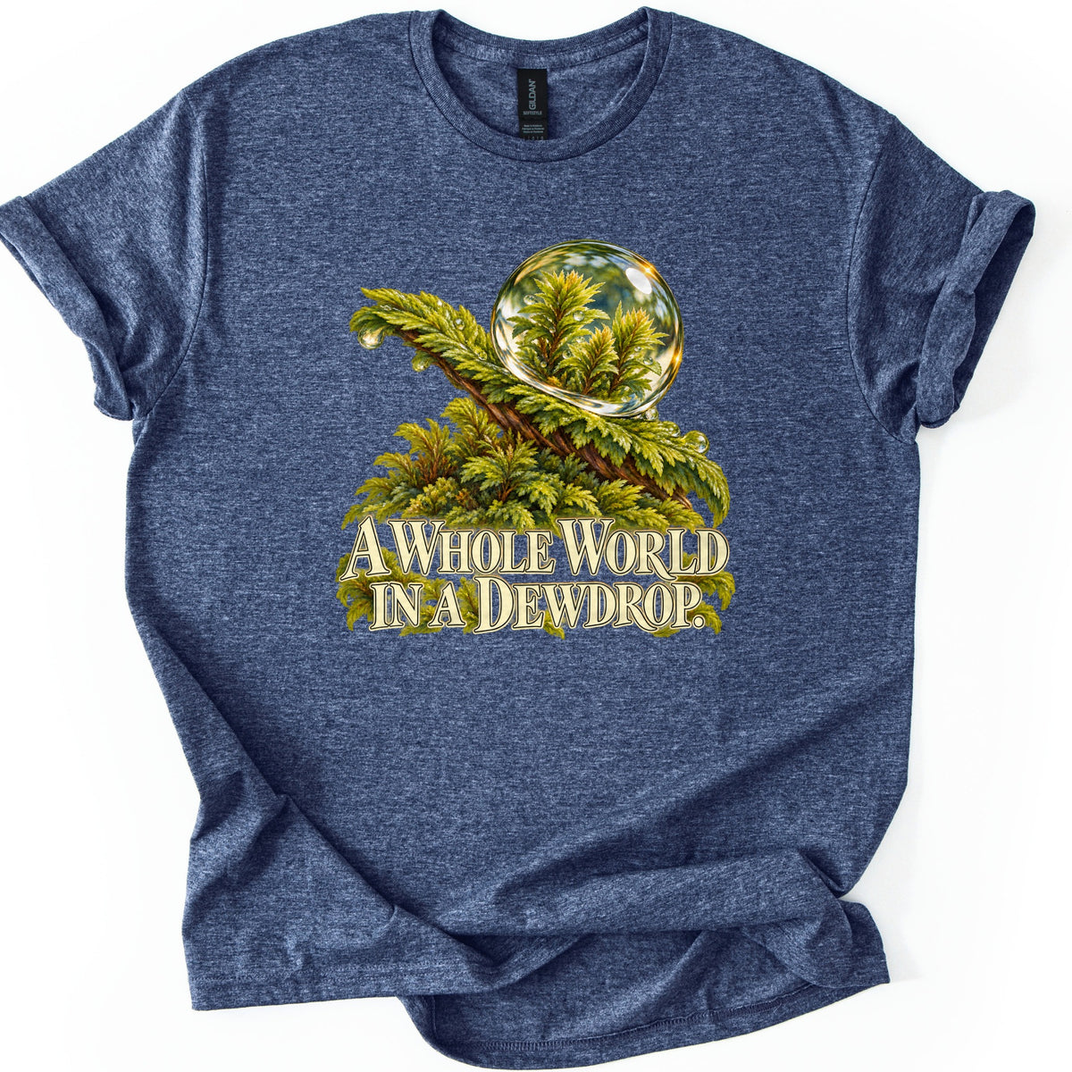 A Whole World in a Dewdrop Comfort Colors T-shirt in Heather Navy, laid flat. Painted green moss holds a glassy dewdrop with miniature plants seen inside it, above the cream words A Whole World in a Dewdrop. The shirt is spread on a white surface with rolled sleeves.