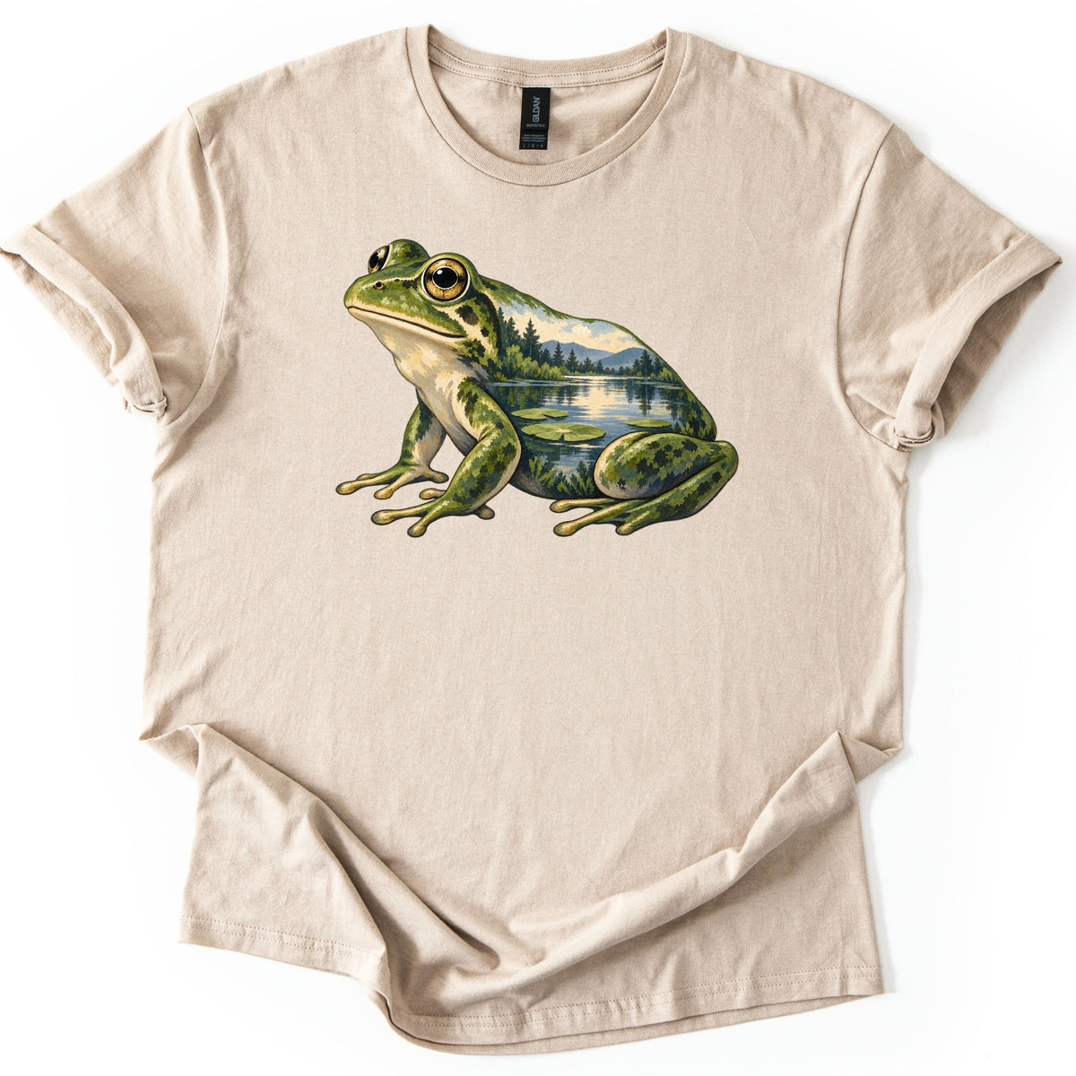 Lily Pond Frog Comfort Colors T-shirt in Sand, laid flat. A painted green frog sits in profile, its body filled with a lake scene of pine trees, mountains and lily pads. No words are printed. The shirt has cuffed sleeves on a white background.