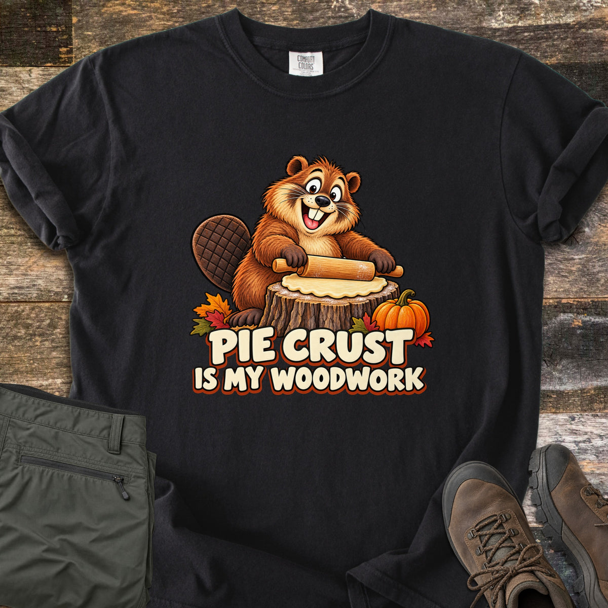 Pie Crust Is My Woodwork Comfort Colors T-shirt in Black, laid flat. A grinning cartoon beaver rolls pie dough on a tree stump, with a small pumpkin and autumn leaves, above the words Pie Crust Is My Woodwork. Olive pants and brown hiking boots sit on a rustic wood surface.