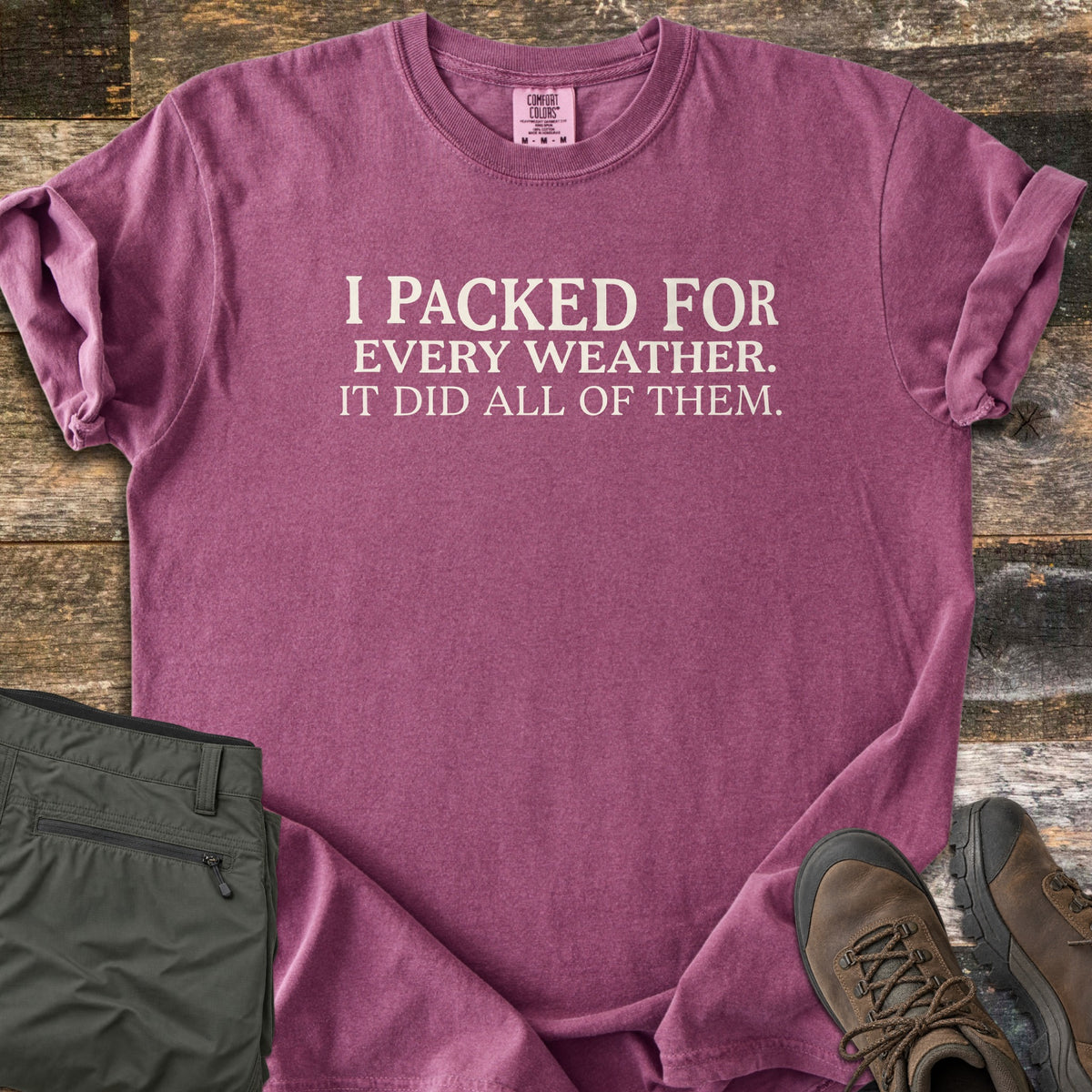 Packed For Every Weather Comfort Colors T-shirt in Berry, laid flat. Cream serif text on the chest reads I PACKED FOR EVERY WEATHER. IT DID ALL OF THEM. The shirt lies on weathered wood planks with folded olive hiking pants and brown hiking boots.