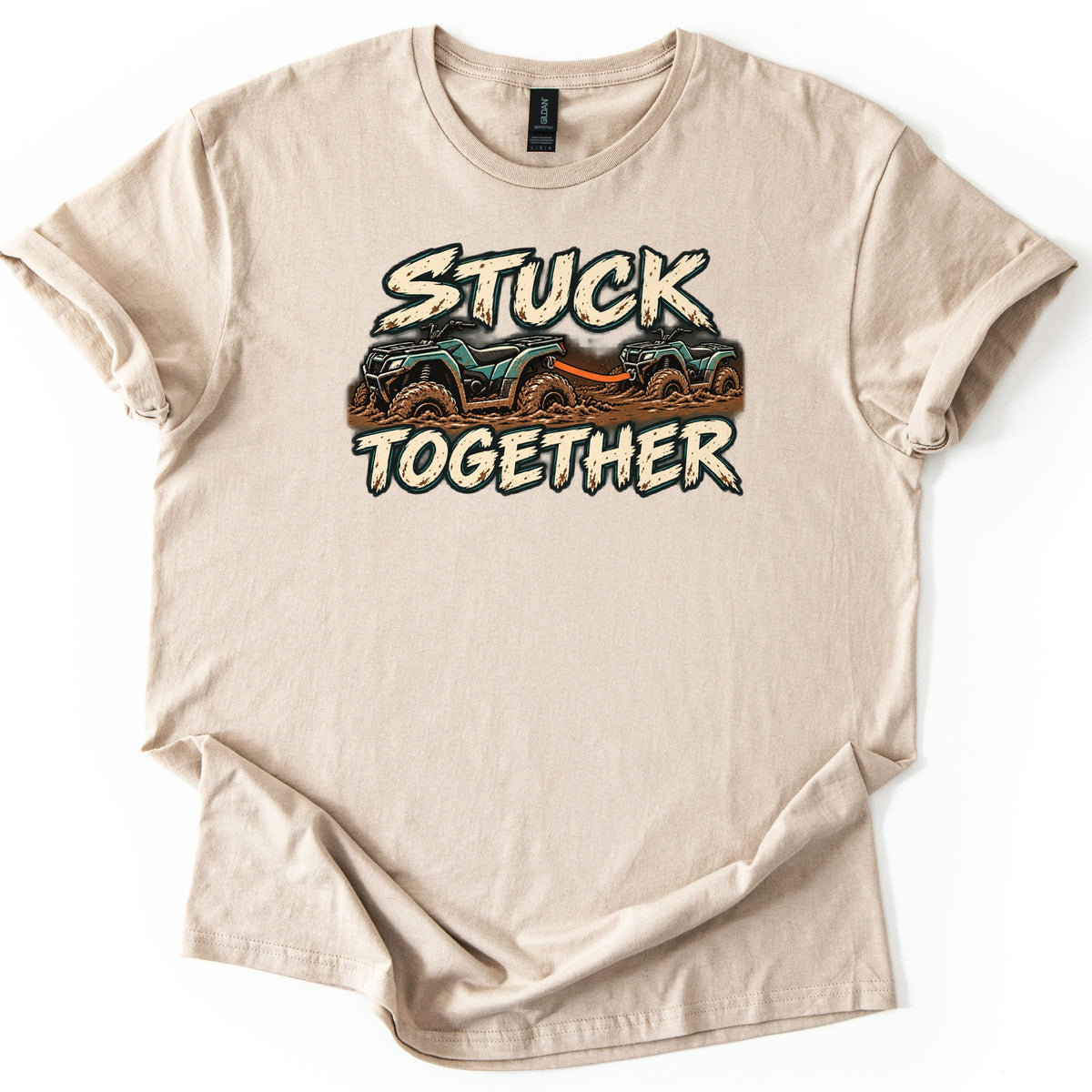 Stuck Together Comfort Colors T-shirt in Sand, laid flat. Two mud-covered green ATVs joined by an orange tow strap sit between the words Stuck Together in distressed cream letters. The shirt has cuffed sleeves and lies on a white background.