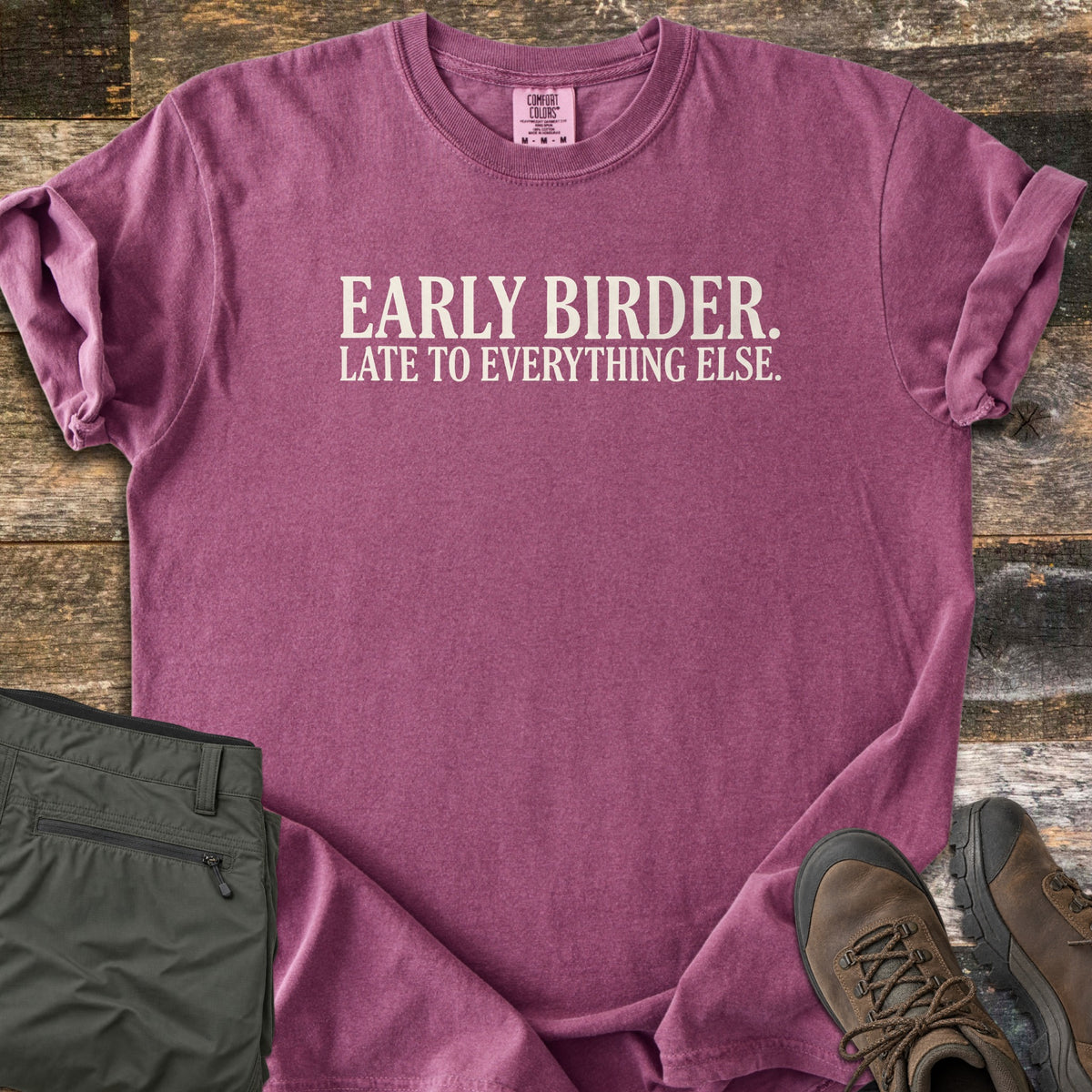 Early Birder Comfort Colors T-shirt in Berry, laid flat. Cream serif text across the chest reads EARLY BIRDER. LATE TO EVERYTHING ELSE. The shirt has rolled sleeves and lies on weathered wood planks beside folded olive hiking pants and brown hiking boots.