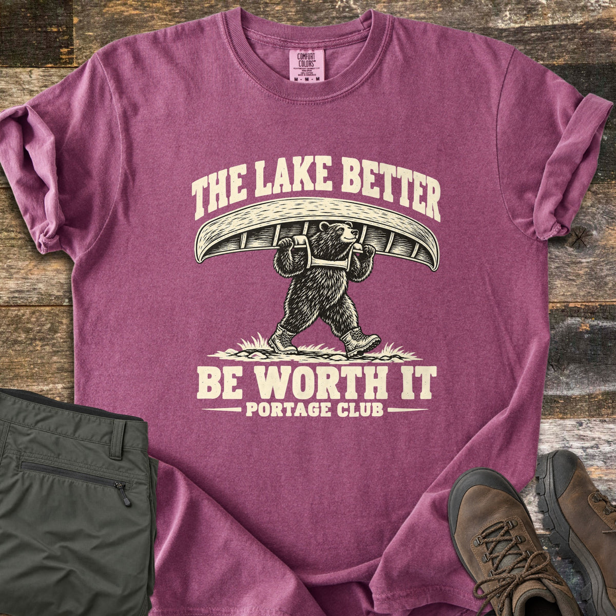 The Lake Better Be Worth It Comfort Colors T-shirt in Berry, laid flat. A bear in hiking boots carries a canoe overhead between the words The Lake Better Be Worth It, with Portage Club below, printed in cream. Shown on weathered wood with folded green pants and brown hiking boots.