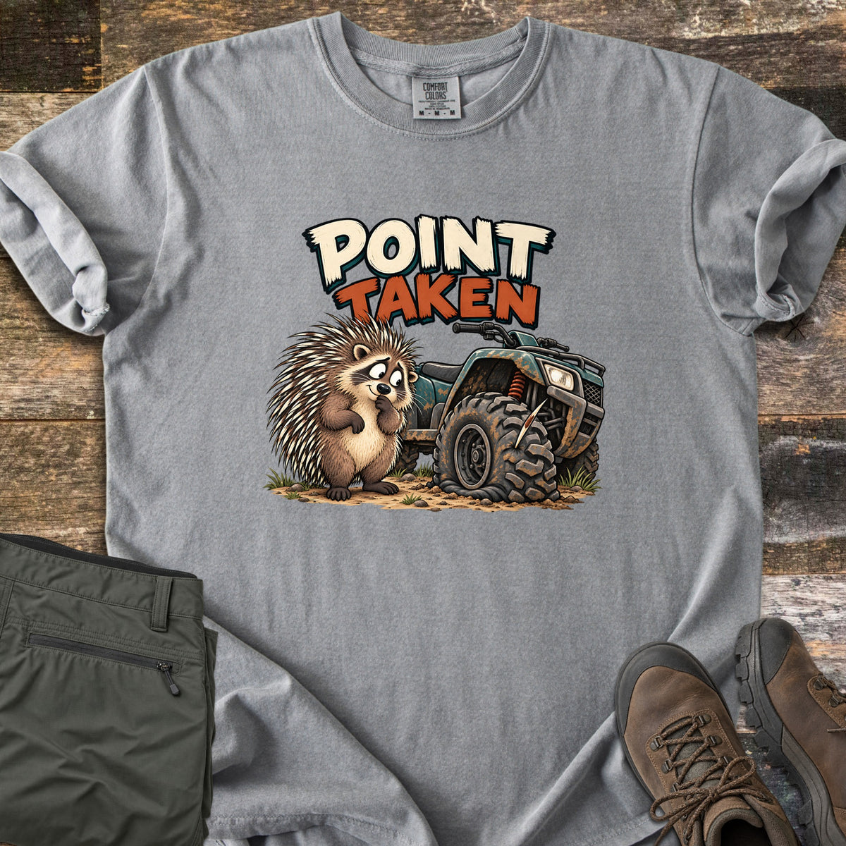 Point Taken Comfort Colors T-shirt in Grey, laid flat. A cartoon porcupine looks sheepishly at a muddy ATV with a flat front tire pierced by a quill, under the words Point Taken. Folded olive pants and brown hiking boots sit beside it on weathered wood.