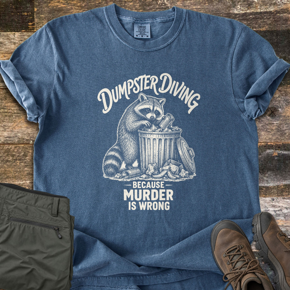Dumpster Diving Because Murder Is Wrong Comfort Colors T-shirt in Denim, laid flat. A cream sketch of a raccoon digging through an overflowing metal trash can, with the words Dumpster Diving above and Because Murder Is Wrong below. Olive cargo pants and brown hiking boots lie on a wood floor.