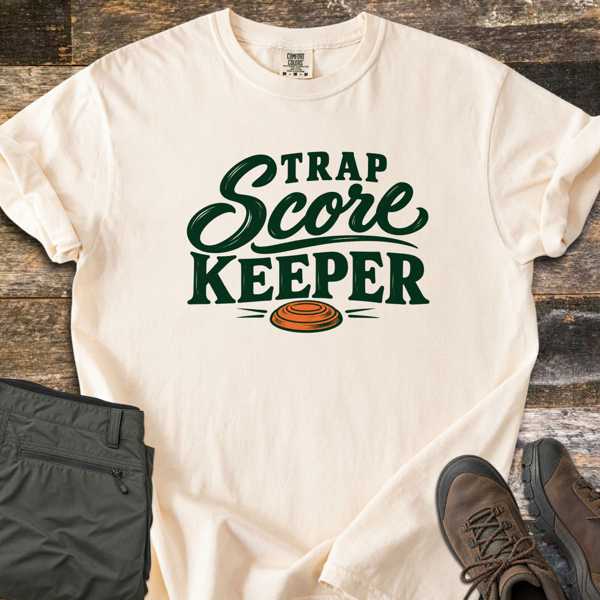 Trap Score Keeper Comfort Colors T-shirt in Ivory, laid flat. Dark green lettering reads Trap Score Keeper above an orange clay target. Sleeves are cuffed on weathered wood, with folded olive cargo pants and brown hiking boots beside it.