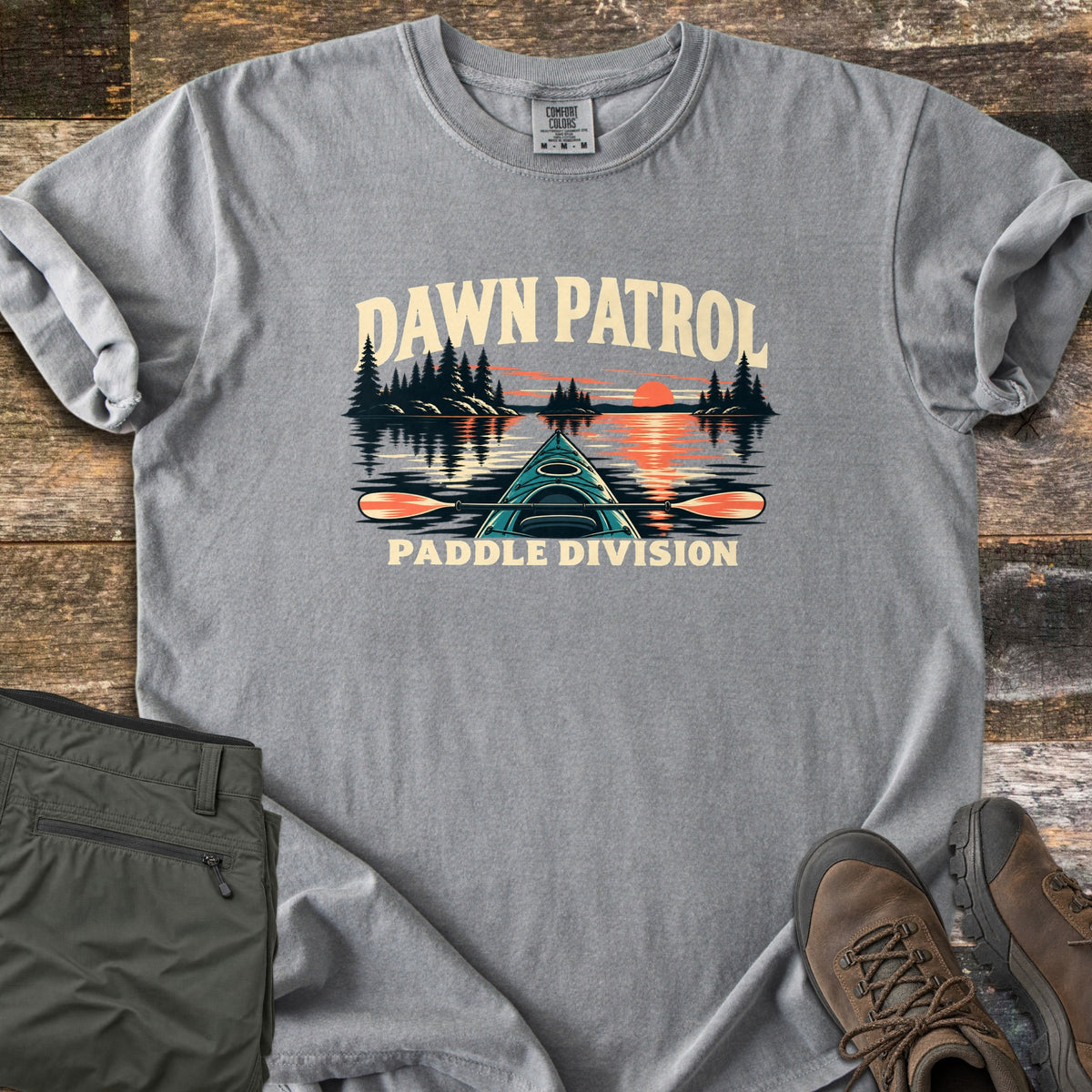 Dawn Patrol Paddle Division Comfort Colors T-shirt in Grey, laid flat. The words Dawn Patrol arch over a teal kayak bow and paddle on a lake with pine islands and an orange sunrise, above the words Paddle Division. Olive hiking pants and brown hiking boots lie beside it on weathered wood.