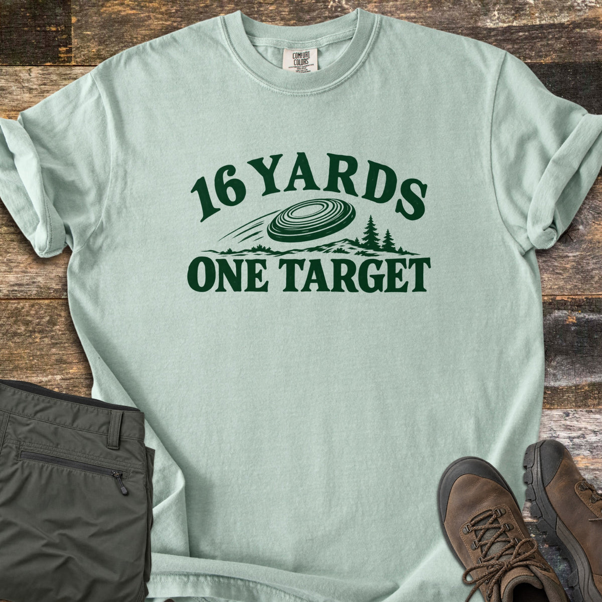 16 Yards One Target Comfort Colors T-shirt in Bay, laid flat. Dark green print of a clay target flying over a hilly treeline with pine trees, between the arched words 16 Yards and One Target. Olive cargo pants and brown hiking boots lie beside it on weathered wood planks.