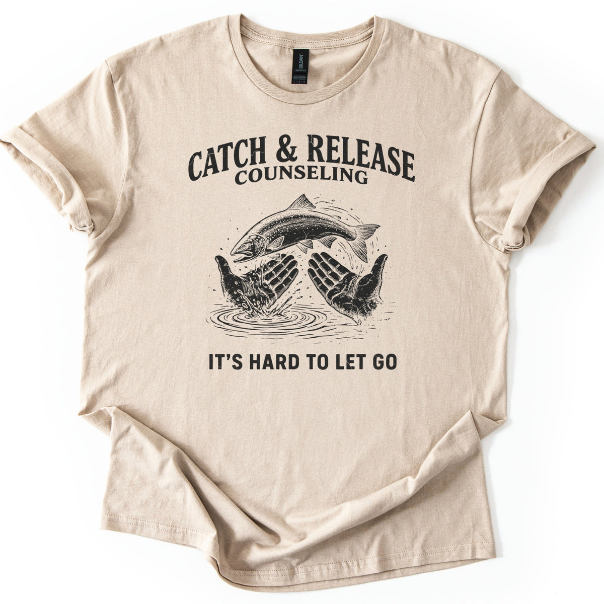 Catch & Release Counseling Comfort Colors T-shirt in Sand, laid flat. Black print reads CATCH & RELEASE COUNSELING above an etched trout leaping from two open hands over rippling water, with IT'S HARD TO LET GO below. Sleeves are cuffed on a white background.
