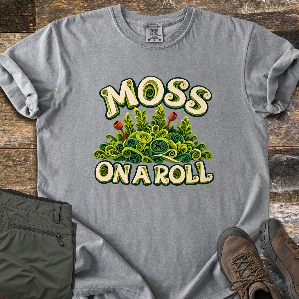 Moss on a Roll Comfort Colors T-shirt in Grey, laid flat. Swirled paper-quill style green moss with two rust-colored sprouts sits between the words Moss On A Roll in cream lettering. Sleeves are cuffed on a wood plank floor beside olive hiking pants and brown hiking boots.