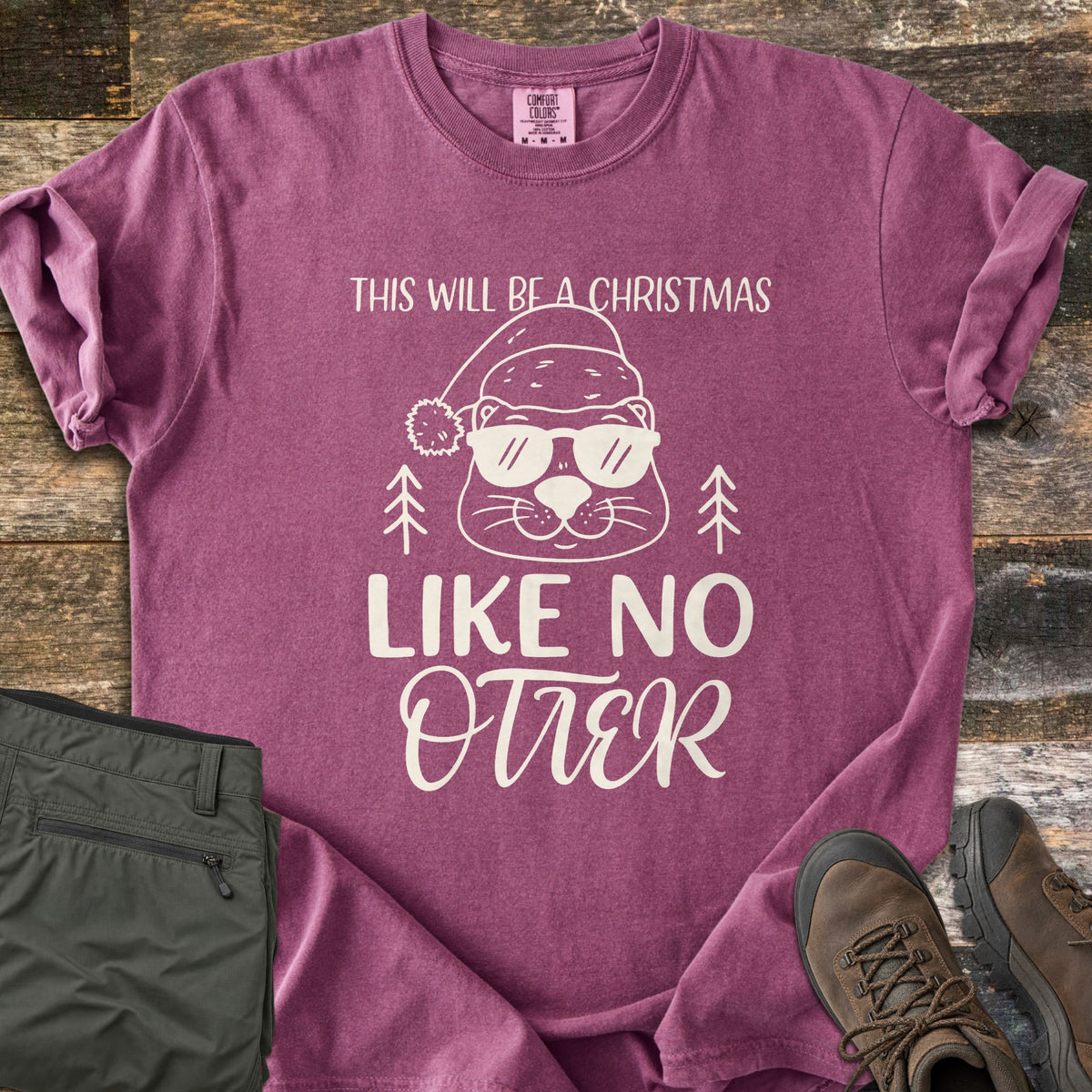 Christmas Like No Otter Comfort Colors T-shirt in Berry, laid flat. A cartoon otter in a Santa hat and sunglasses sits between two pine trees, with This Will Be a Christmas above and Like No Otter below, in cream. Olive cargo pants and brown hiking boots lie on weathered wood.