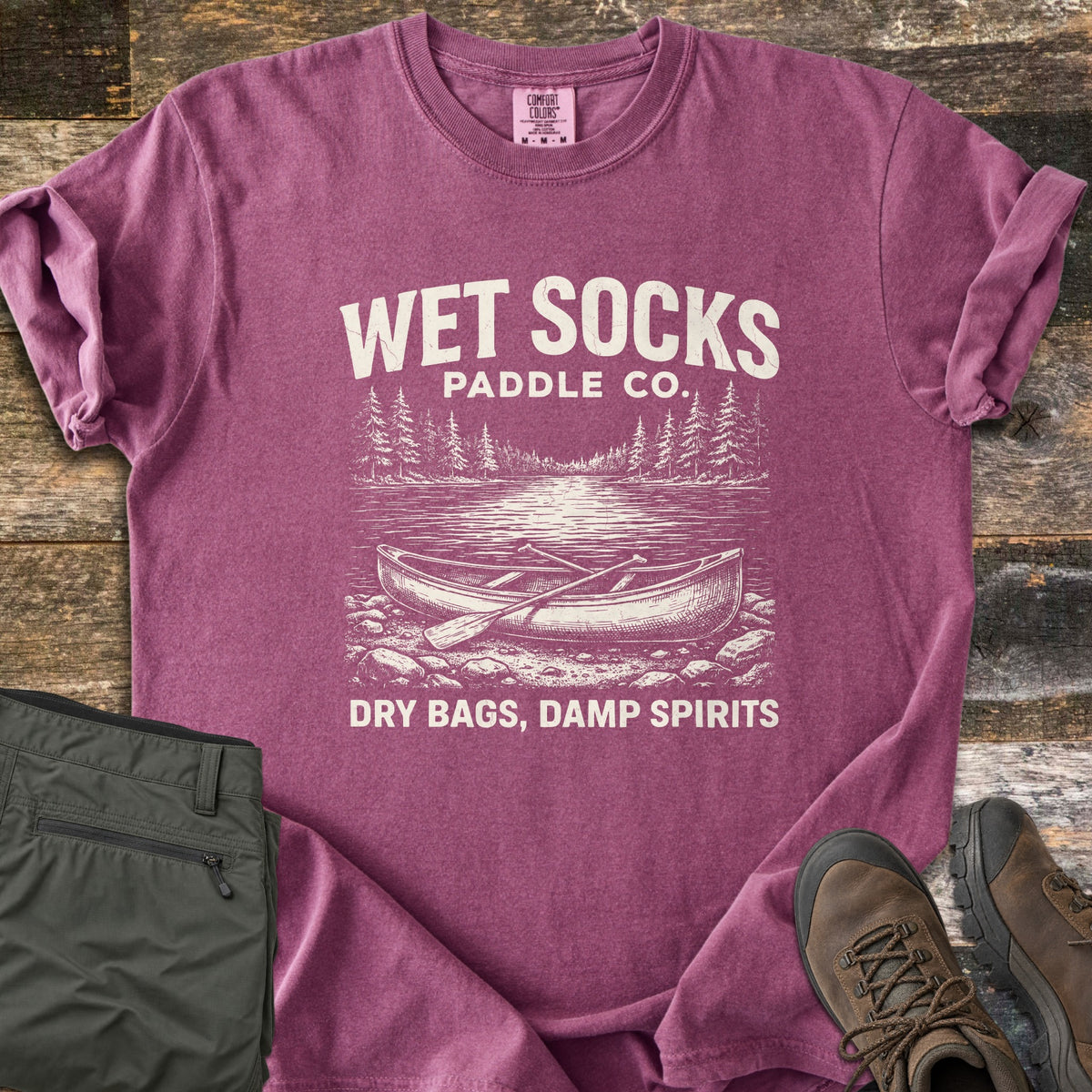 Wet Socks Paddle Co Comfort Colors T-shirt in Berry, laid flat. Cream print reads Wet Socks Paddle Co. above a sketched canoe and paddle on a rocky lakeshore with pines, then Dry Bags, Damp Spirits. Olive hiking pants and brown boots sit on weathered wood.