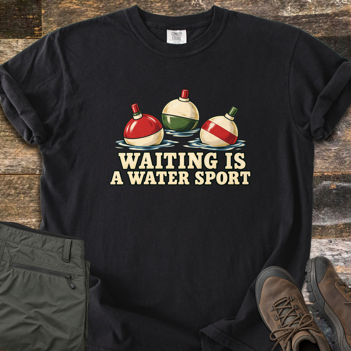Waiting Is a Water Sport Comfort Colors T-shirt in Black, laid flat. Three red, green and cream fishing bobbers float in water above the words Waiting Is a Water Sport. The shirt lies on weathered wood beside folded olive pants and brown hiking boots.
