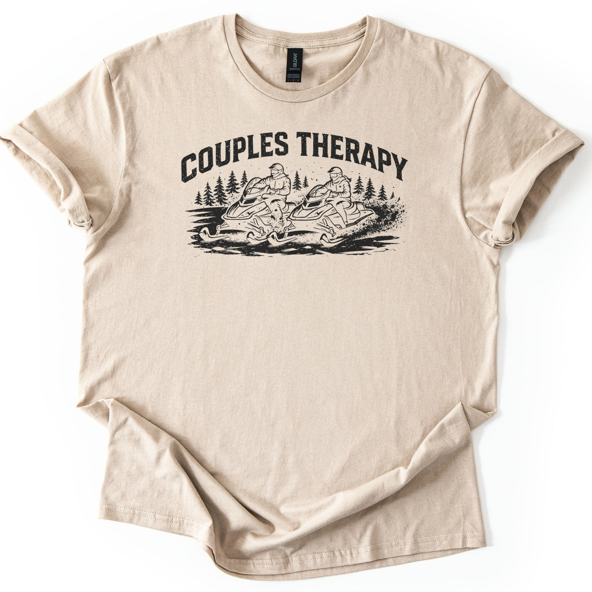 Couples Therapy Snowmobile Comfort Colors T-shirt in Sand, laid flat. Black print of the arched words Couples Therapy above two helmeted riders on snowmobiles racing side by side through snow with pine trees behind. Sleeves are cuffed on a white background.