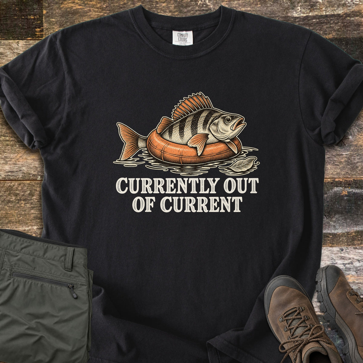 Currently Out of Current Comfort Colors T-shirt in Black, laid flat. A striped perch lounges on an orange inner tube floating in water beside a leaf, above the words Currently Out of Current in cream lettering. Olive hiking pants and brown hiking boots sit on weathered wood planks.