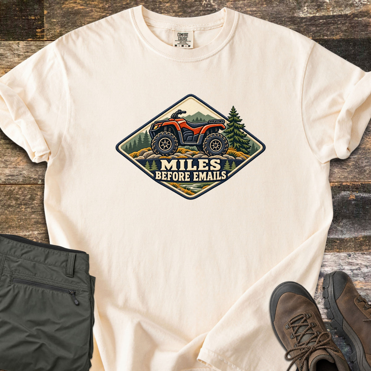 Miles Before Emails Comfort Colors T-shirt in Ivory, laid flat. A diamond-shaped patch shows a red ATV on rocks with pine trees and mountains above the words Miles Before Emails. Sleeves rolled, on weathered wood with green pants and brown hiking boots.