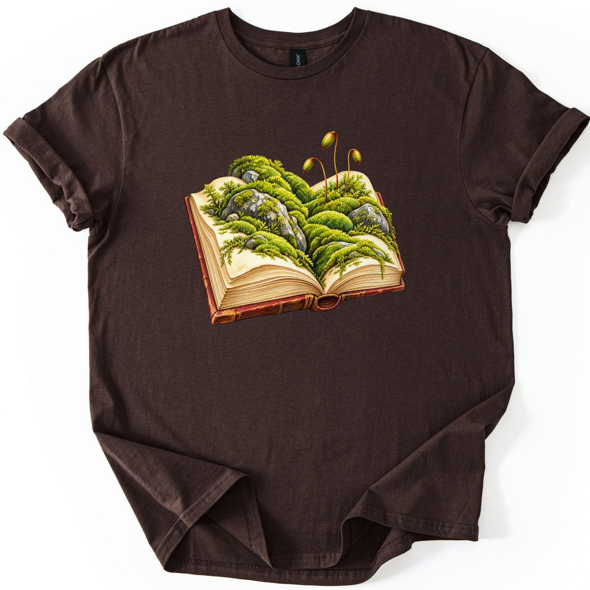 Moss Storybook Comfort Colors T-shirt in Dark Chocolate, laid flat. An open vintage book with a red-brown leather cover is overgrown with green moss and mossy gray rocks, and three spore stalks with small capsules rise above. The shirt lies on a white background with rolled sleeves.