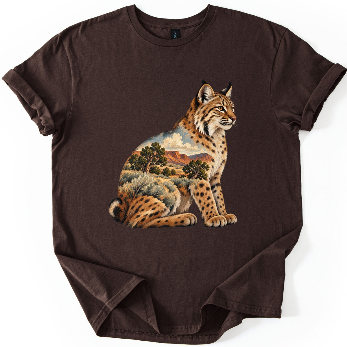 Bobcat Sagebrush Canyon Comfort Colors T-shirt in Dark Chocolate, laid flat. A painted bobcat sits facing right, its body filled with a desert scene of red canyon cliffs, green trees, sagebrush and clouds. No words are printed. The shirt lies on a white surface with cuffed sleeves.