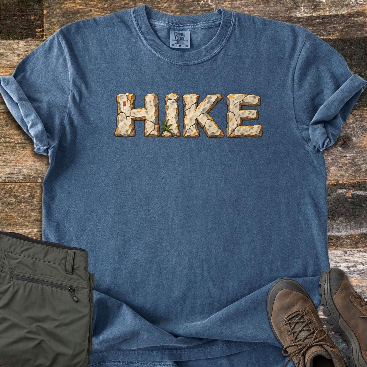 Hike Comfort Colors T-shirt in Denim, laid flat. The word HIKE is printed across the chest in cracked tan stone letters with orange edges, a small fern growing at the base of the I. Olive hiking pants and brown leather boots lie beside it on weathered wood.