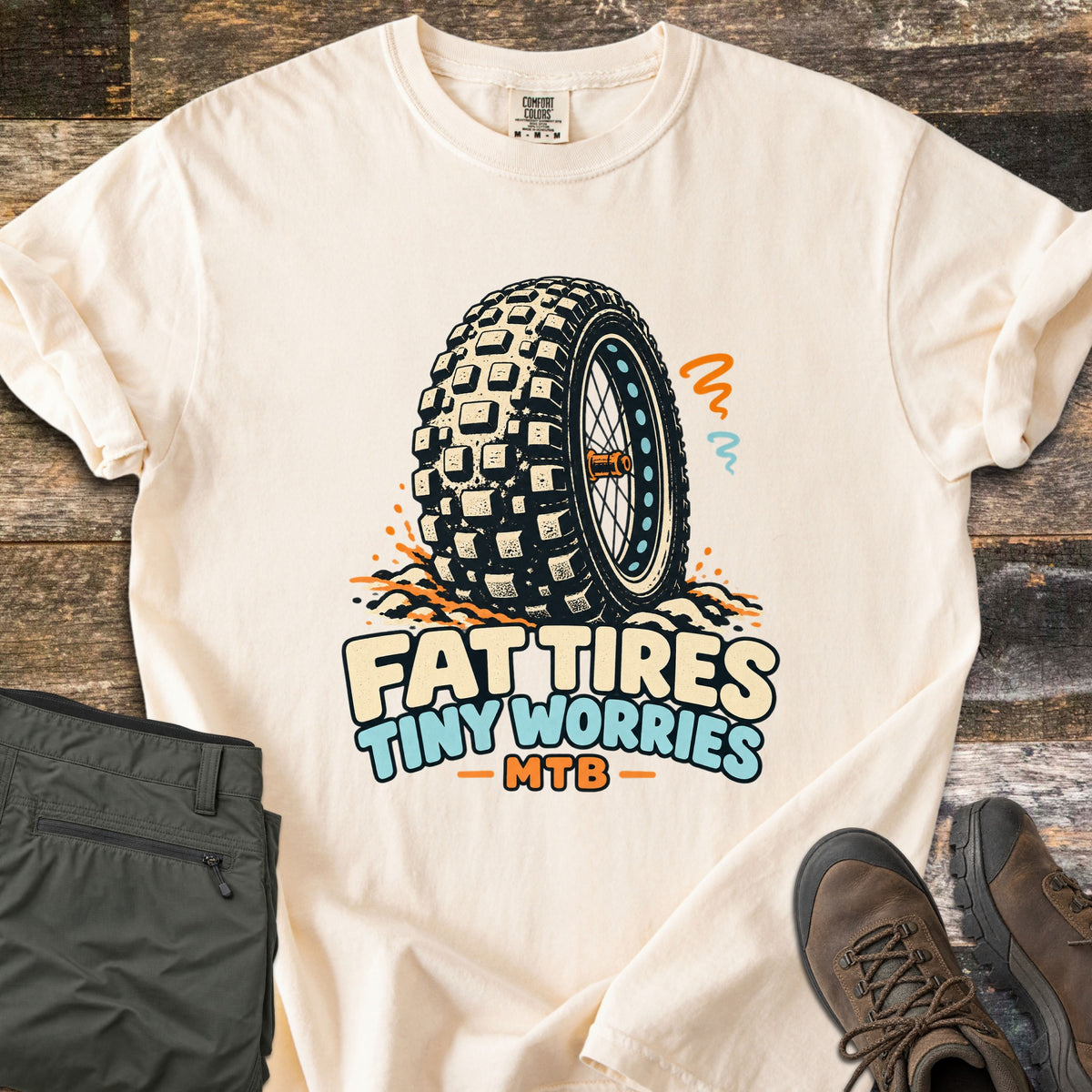 Fat Tires Tiny Worries Comfort Colors T-shirt in Ivory, laid flat. A knobby fat bike tire kicking up dirt, with orange and blue squiggles, above the words Fat Tires Tiny Worries MTB. Sleeves rolled, on weathered wood with olive pants and brown hiking boots.
