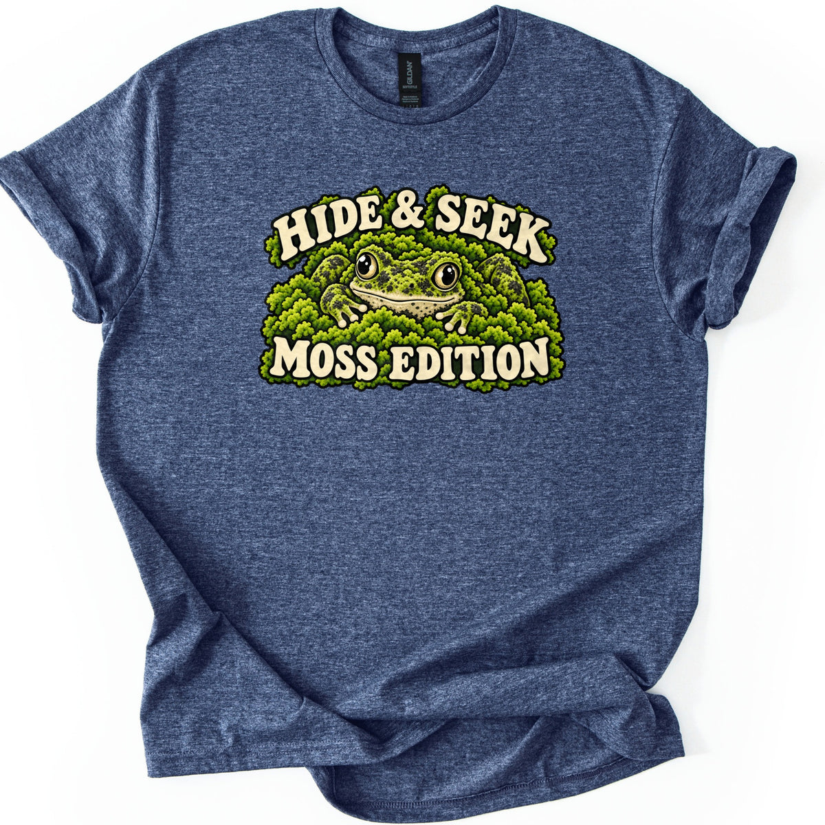 Hide & Seek Moss Edition Comfort Colors T-shirt in Heather Navy, laid flat. A mottled green frog peers out from clumps of green moss under the arched cream words Hide & Seek, with Moss Edition beneath. The shirt is shown on a white background with cuffed sleeves.