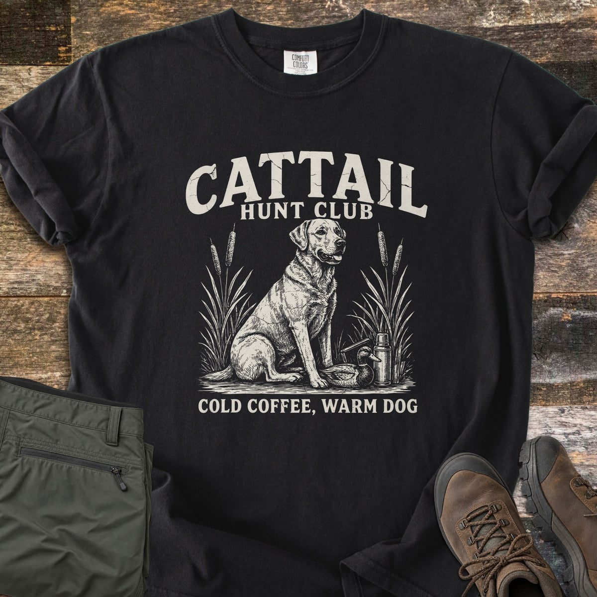 Cattail Hunt Club Comfort Colors T-shirt in Black, laid flat. Cream print of a Labrador sitting among cattails beside a duck decoy and thermos, under the words Cattail Hunt Club, above Cold Coffee, Warm Dog. On weathered wood with olive pants and brown hiking boots.